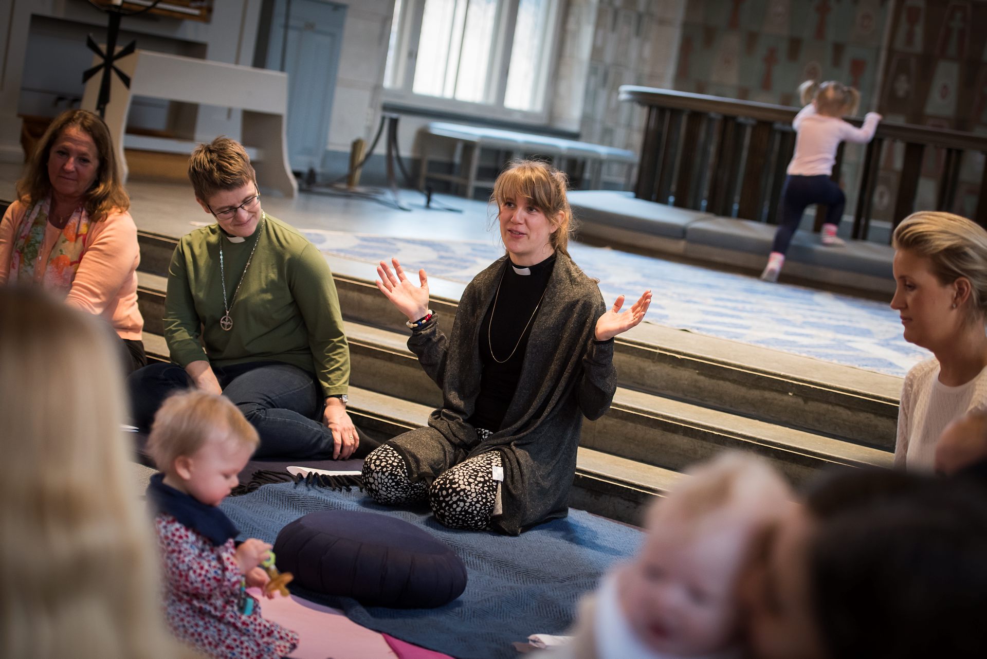 På en blå filt på kyrkgolvet nedanför koret sitter en kvinnlig präst. Vid hennes vänstra sida sitter en kvinnlig diakon. I bakgrunden kan man se en liten flicka på väg att klättra på knäfallet framför altaret. på bilden syns även fler små barn och deras mammor.