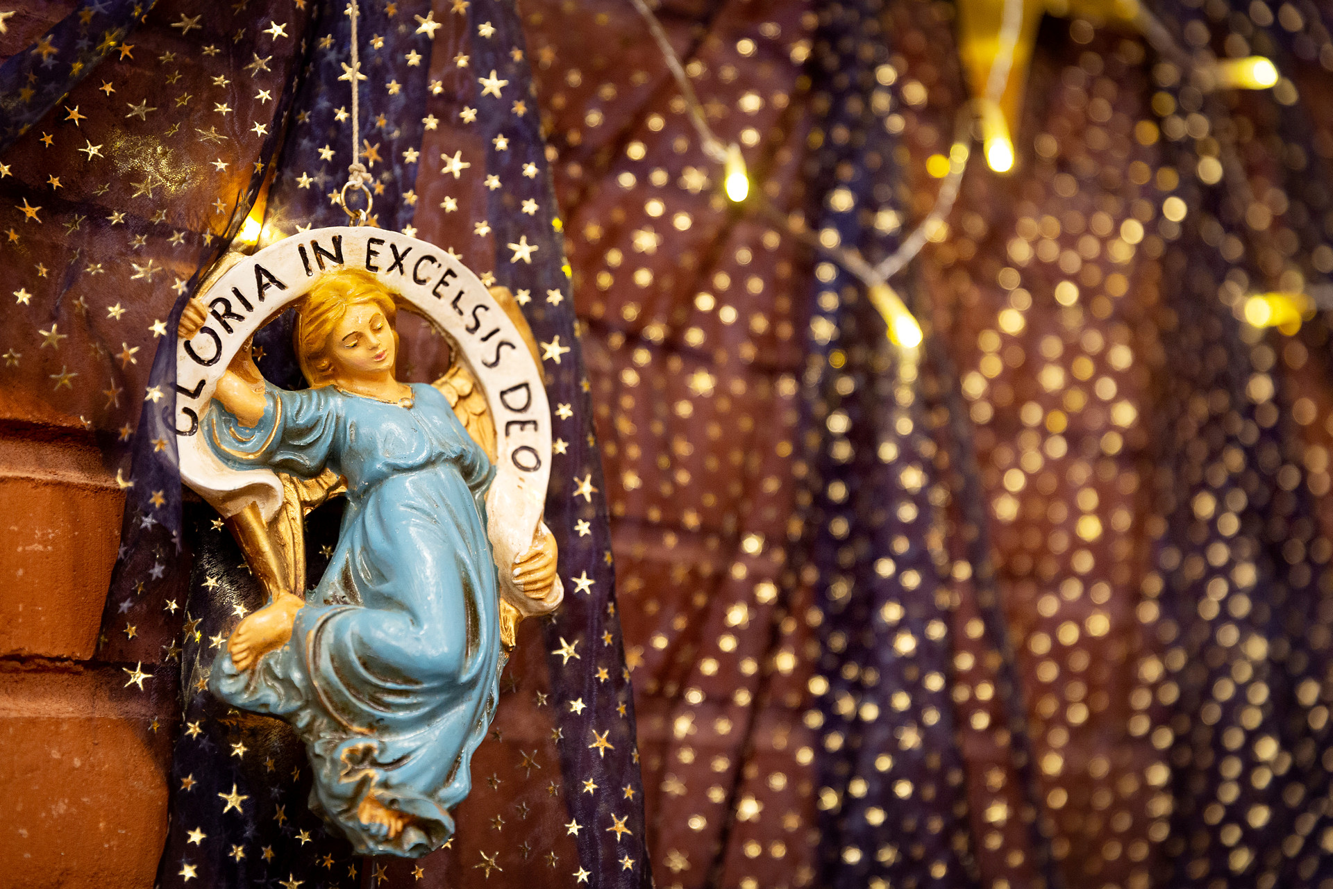 En prydnadsängel med ljusblå klänning och blont hår hänger framför ett genomskinligt och svart draperi med guldstjärnor på. Över ängel står texten "Gloria in excelsis deo" på en banderoll som ängel håller i.