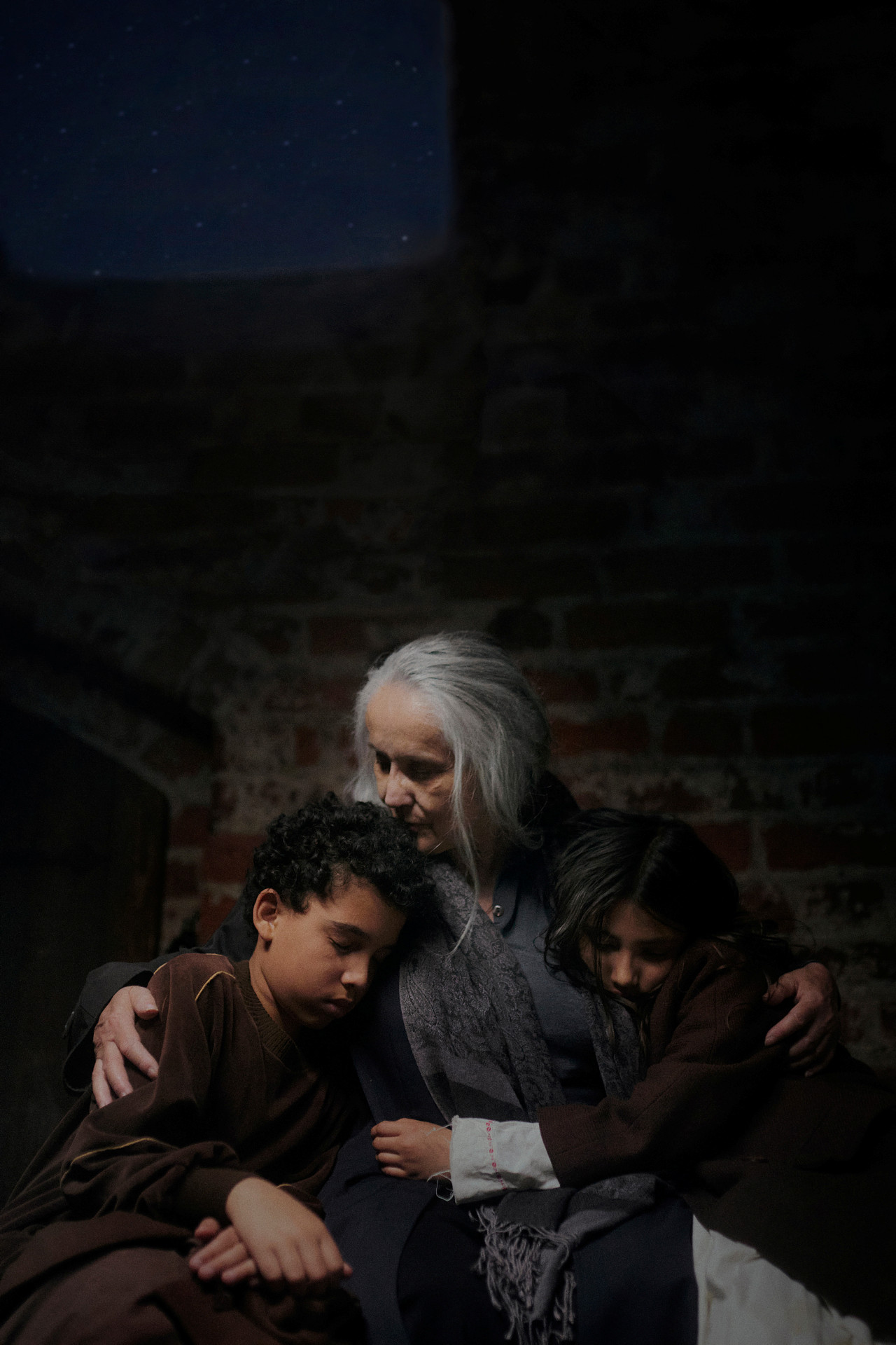 En gråhårig äldre dam håller om sina barnbarn, en pojke och en flicka, i ett mörkt rum i krigstid.