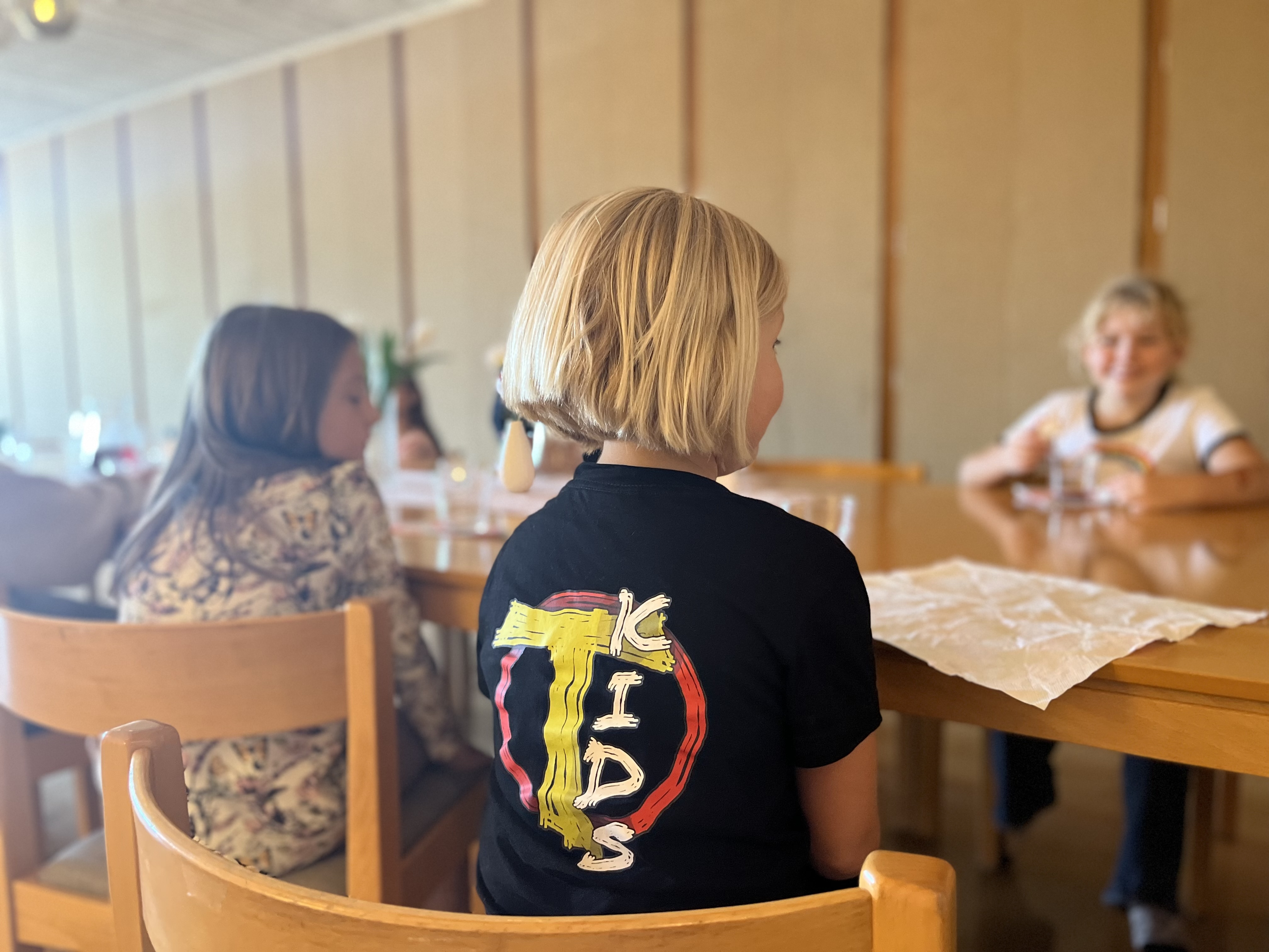 En blond flicka sitter med ryggen vänd mot kameran med en t-shirt som det står "T-kids" på.