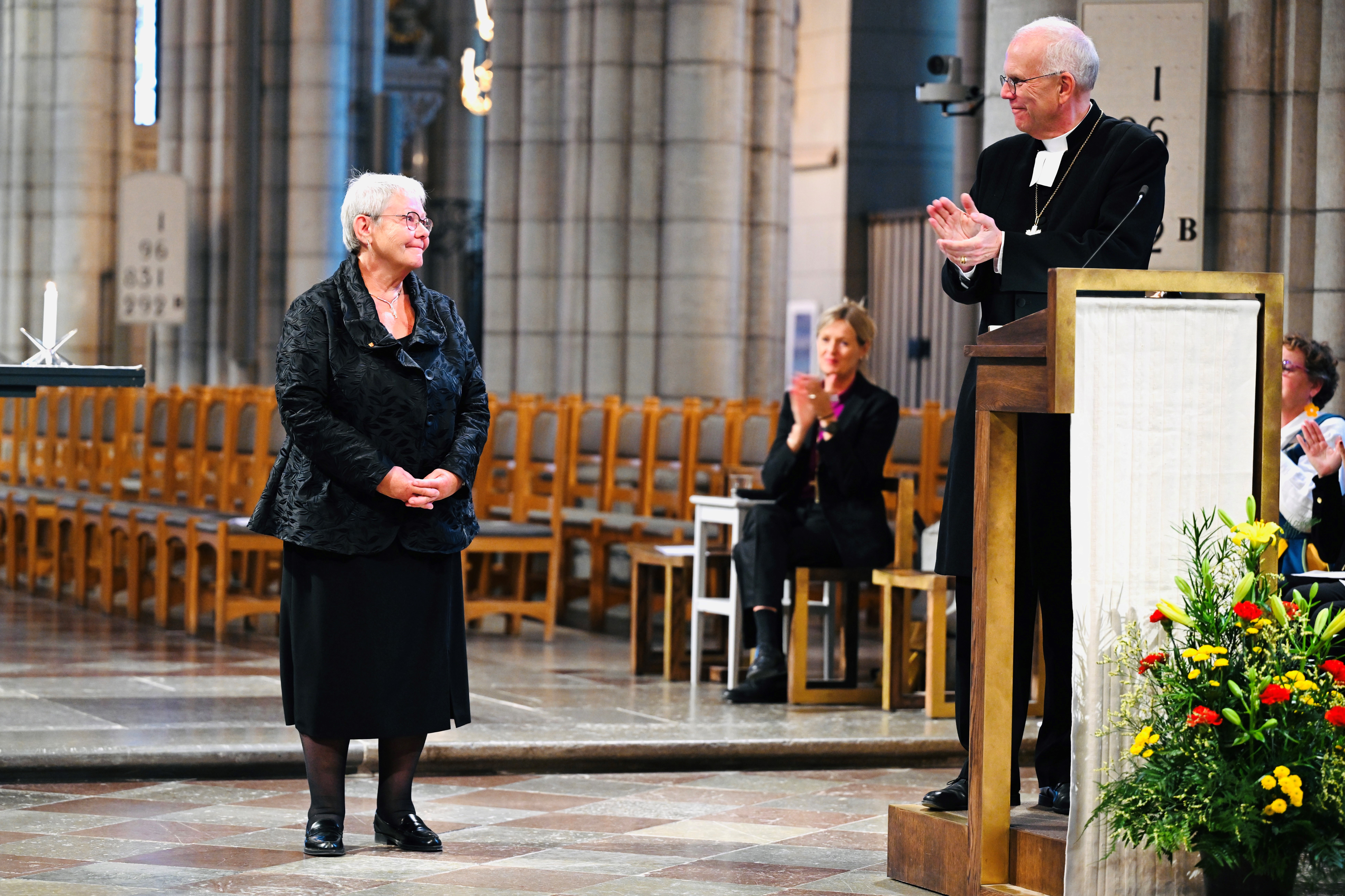 Svartklädd kvinna står framför ärkebiskopen som klappar händerna.