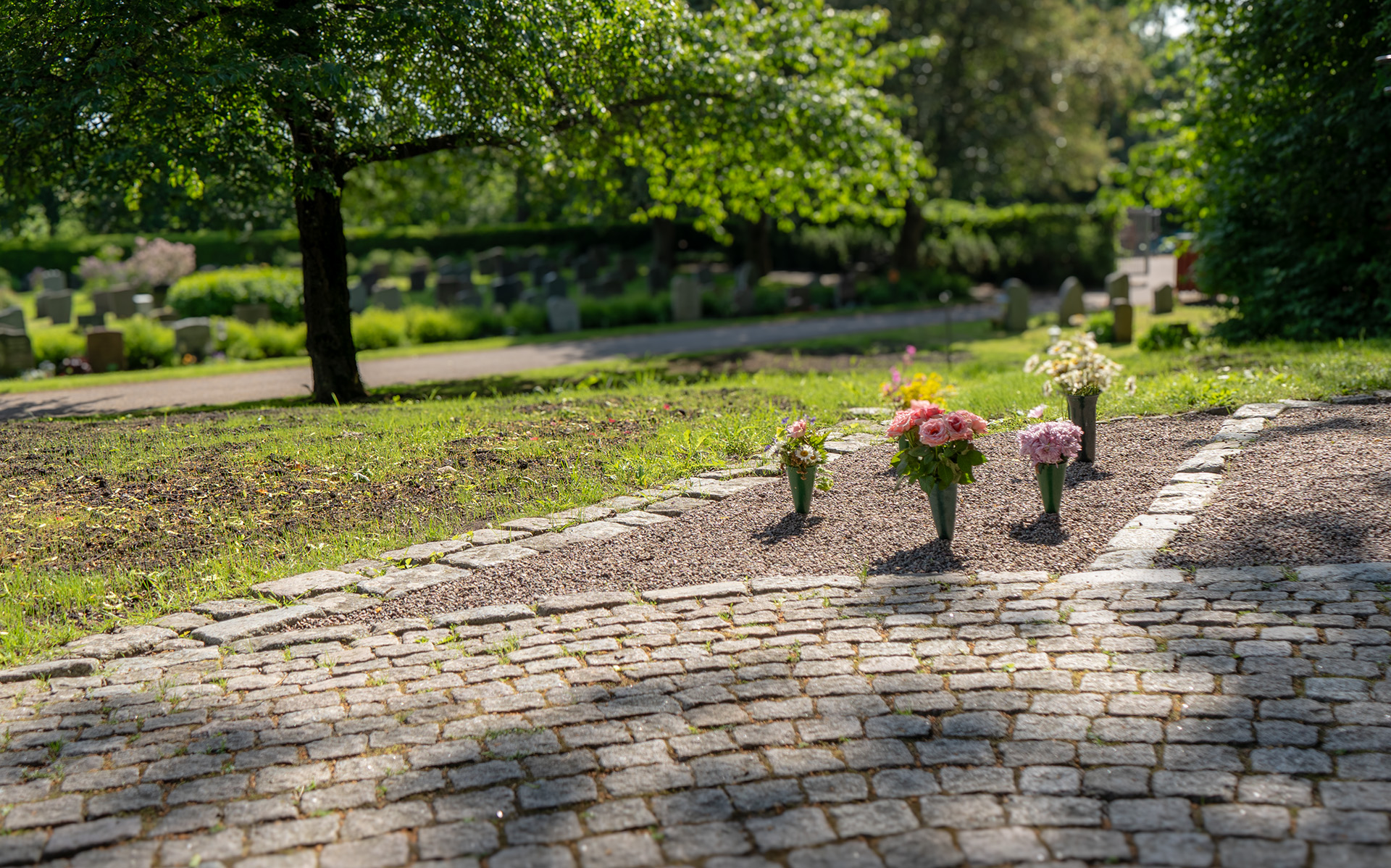 Halvcirkel av stenar framför två grusytor med nedstuckna vaser och snittblommor i en minneslund. Intill finns gräs och i bakgrunden syns delar av kyrkogården.