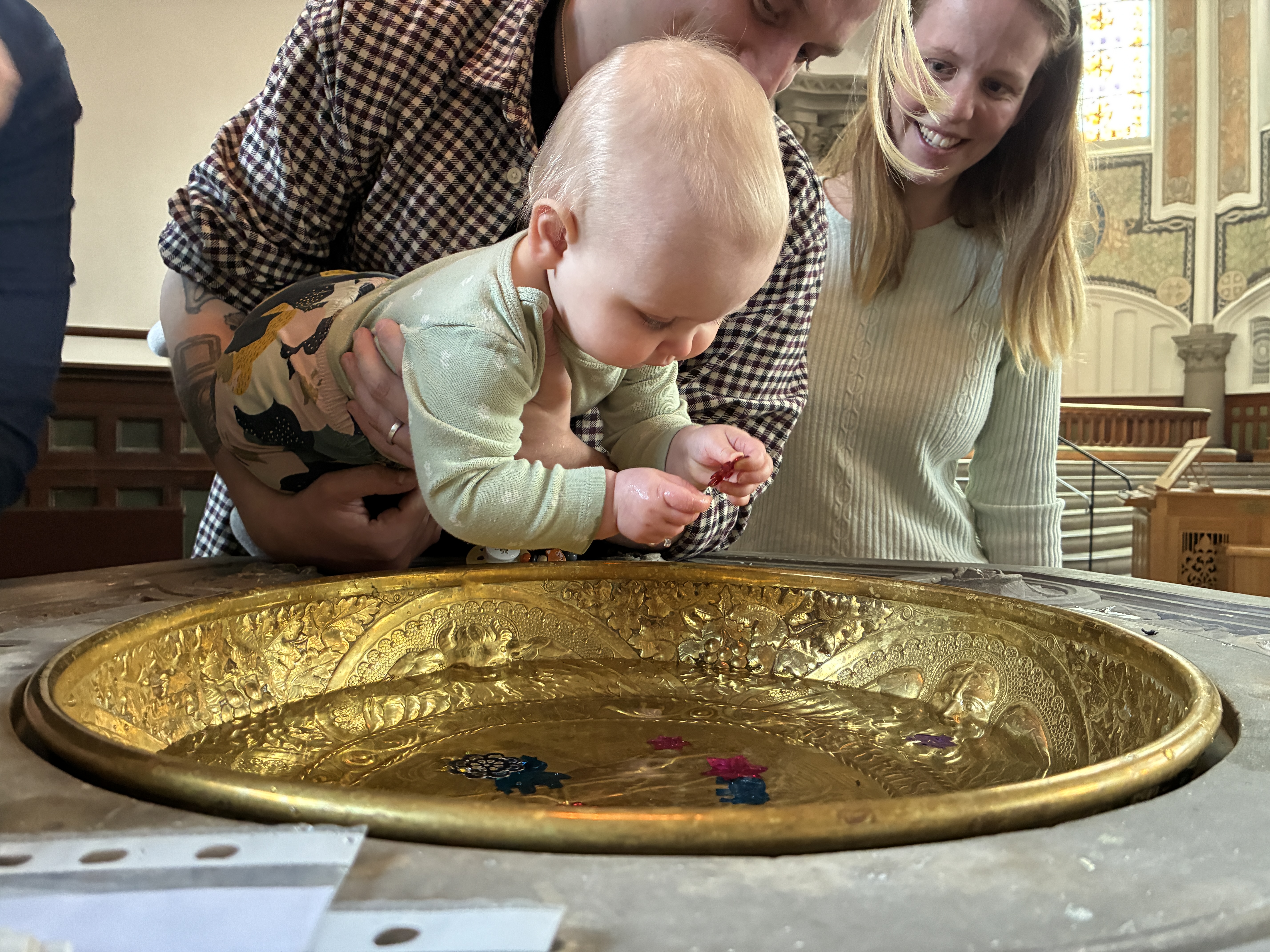 Bebis hålls av en vuxen över dopfunten med en röd paljett i handen