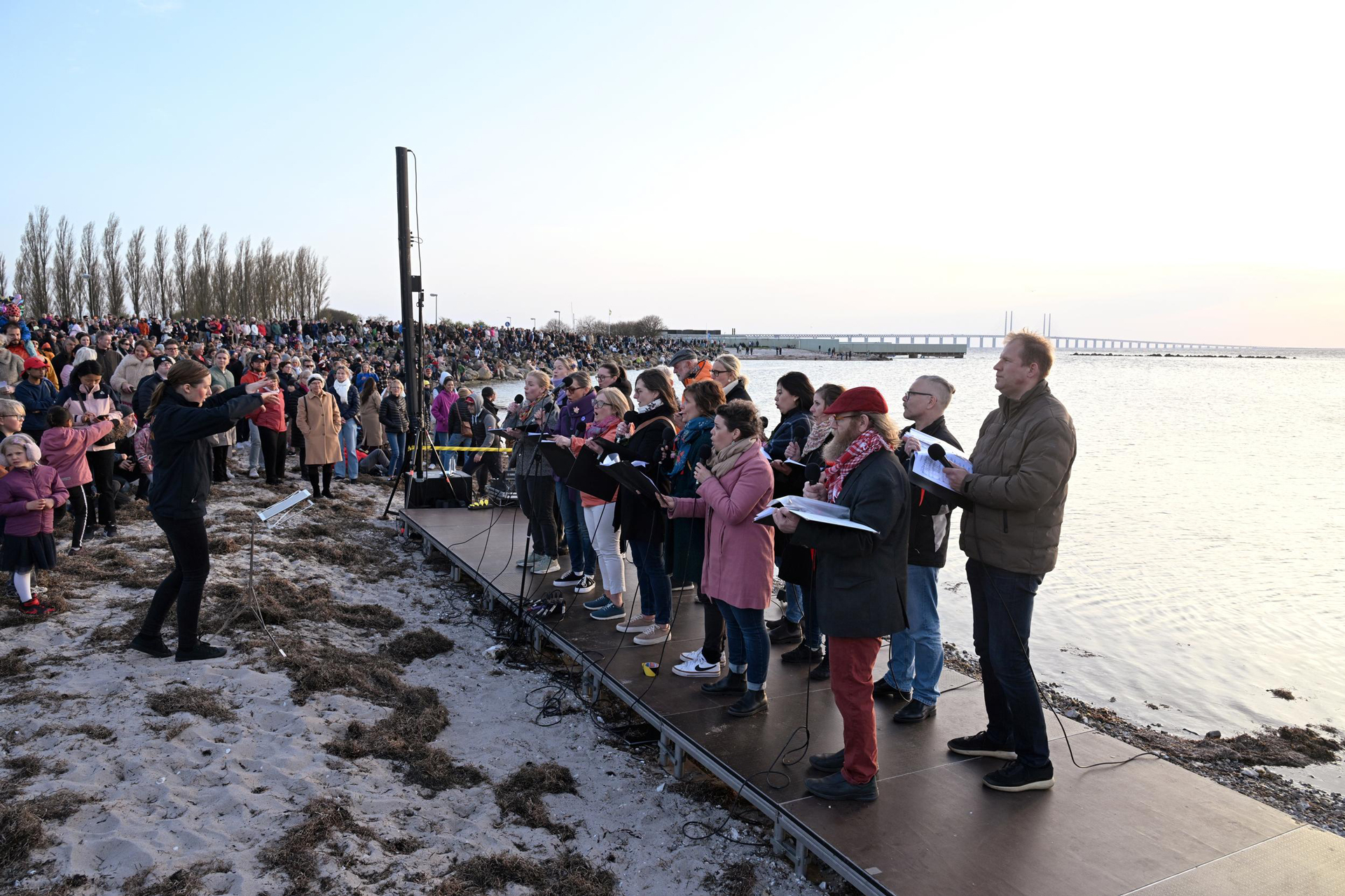En grupp människor sjunger utomhus vid hav och Öresundsbron.