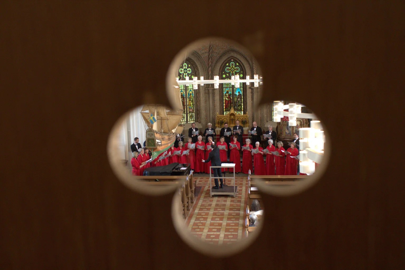 Sjungande kör stående framme i koret på en kyrka. Främre raden är klädda i röda kåpor och bakre raden är klädda i svart. Fotot är taget genom en fyrklöver-formad hålighet från en läktare.