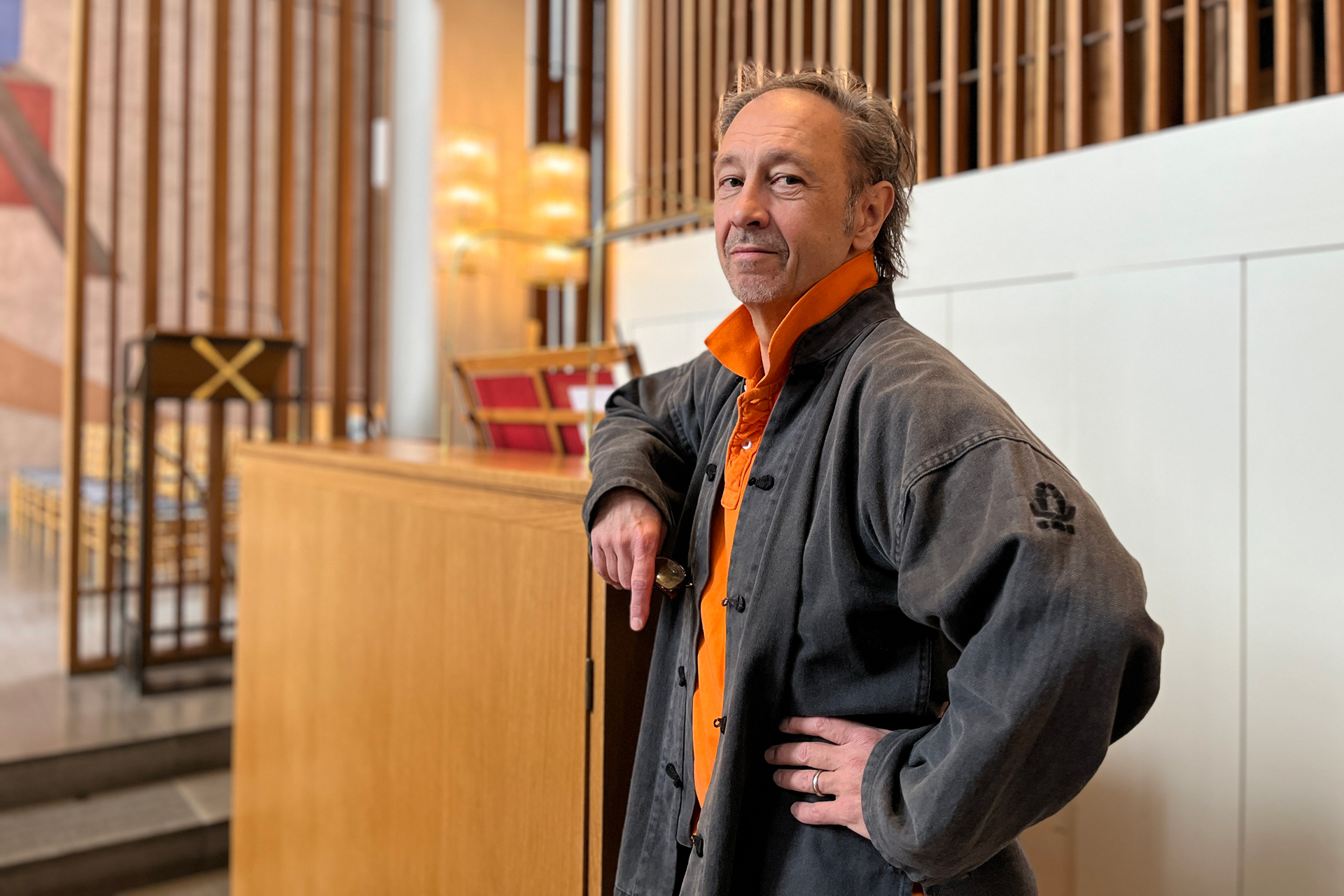 Man med gråsprängt hår klädd i orange skjorta och svart jacka i kyrkorum.