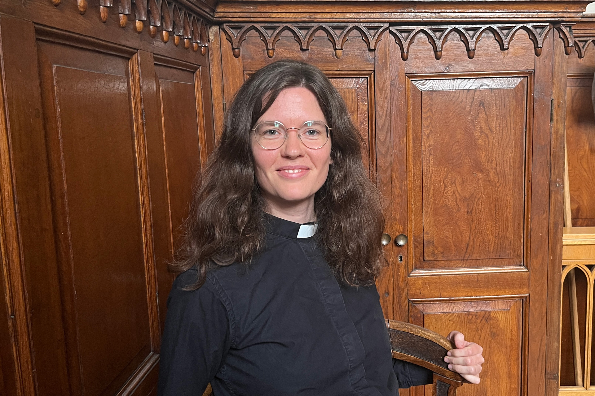 Närbild på yngre kvinna med långt brunt hår och glasögon klädd o svart prästskjorta mot träfond.
