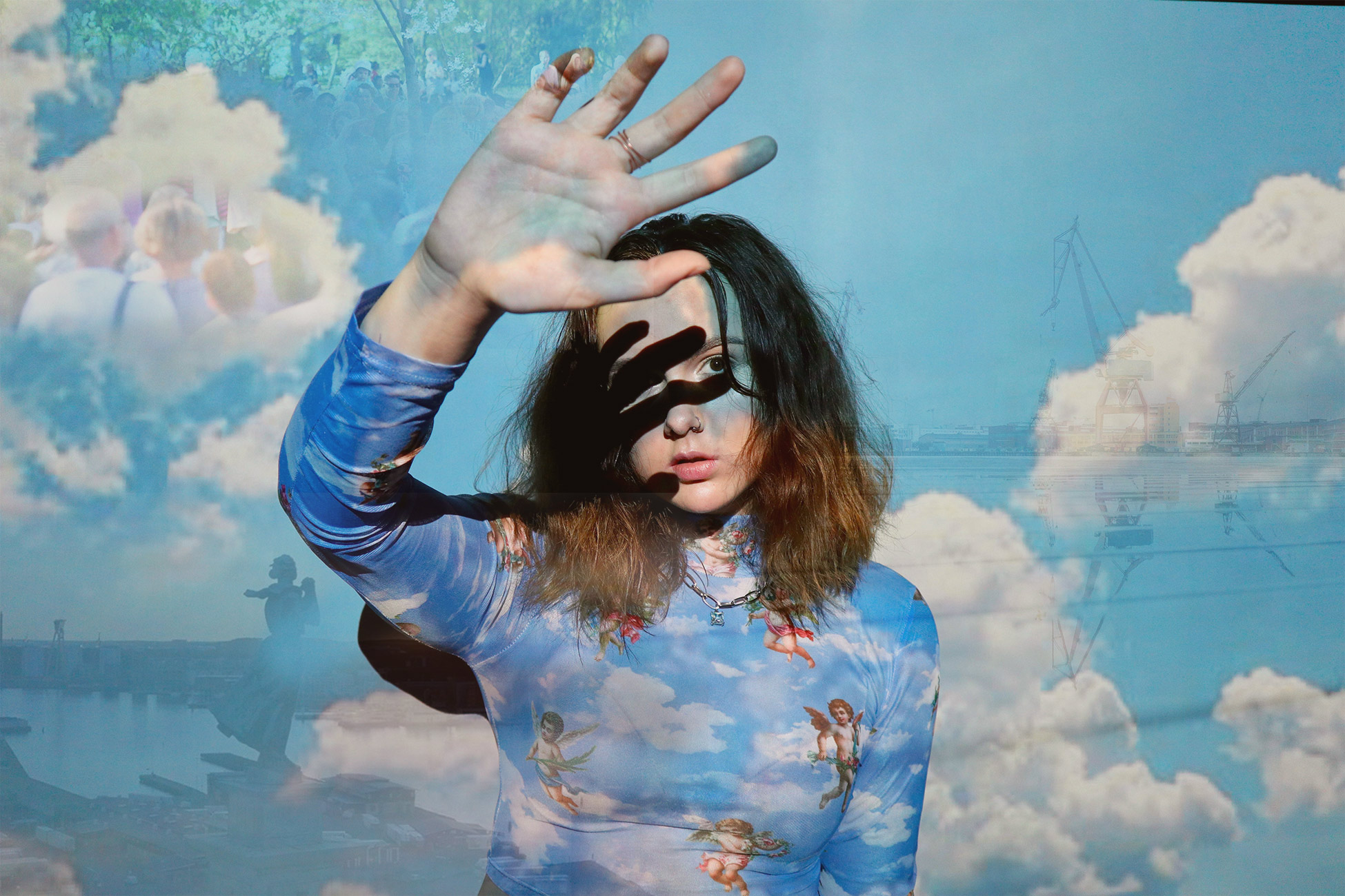 Ung kvinna skuggar ansiktet med handen mot starkt ljussken. Dubbelexponerad bild med himmel och Göteborgs skyline med kranar Sjömanshustrun. 