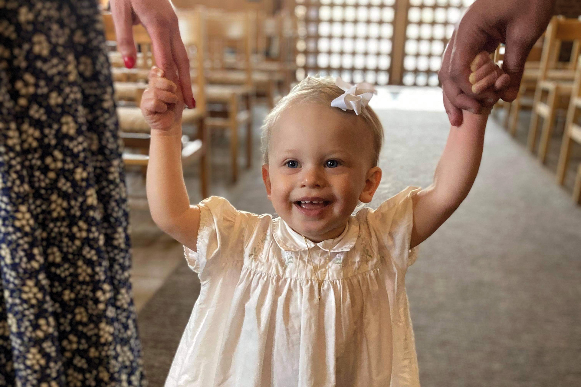 En glad flicka i vit klänning håller två vuxna i varsin hand.