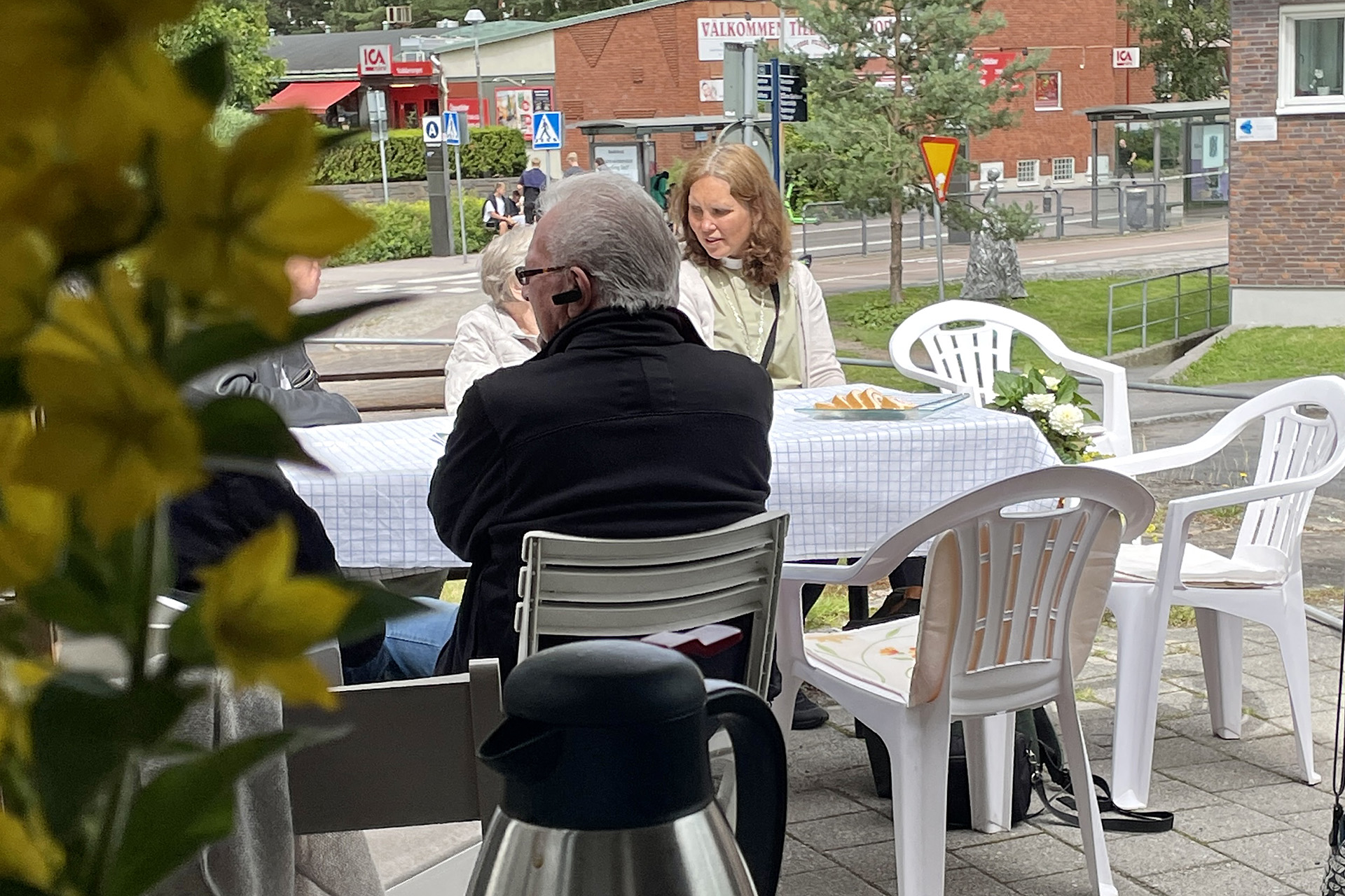 Personer sitter utomhus vid bord och fikar. I förgrunden syns en blomma, i bakgrunden en mataffär.