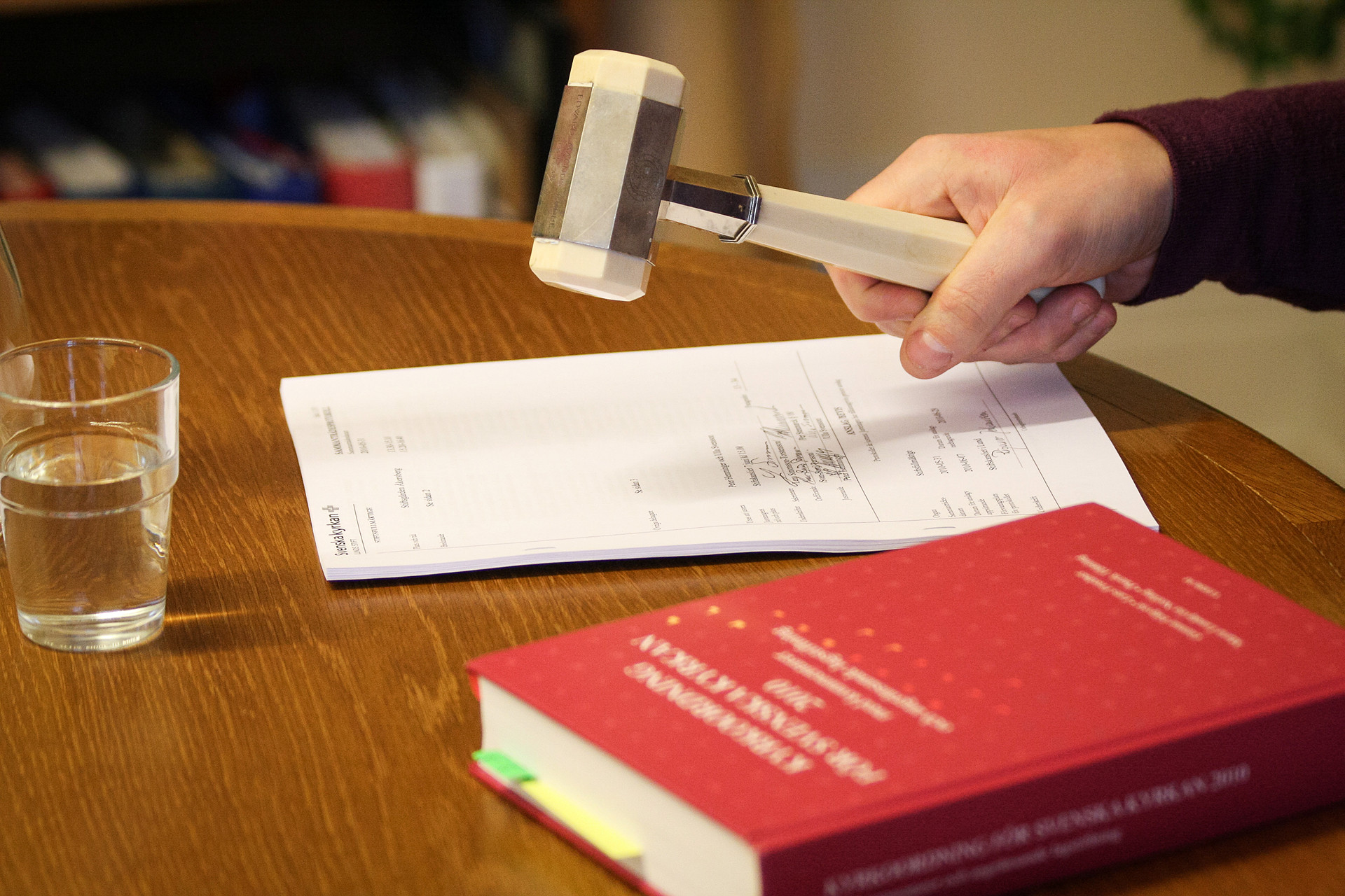 En mörkbrun bordsyta. På det står ett vattenglas, där ligger också ett protokoll samt en tjock röd bok. Någon är på väg att slå en ordförandeklubba i bordet.