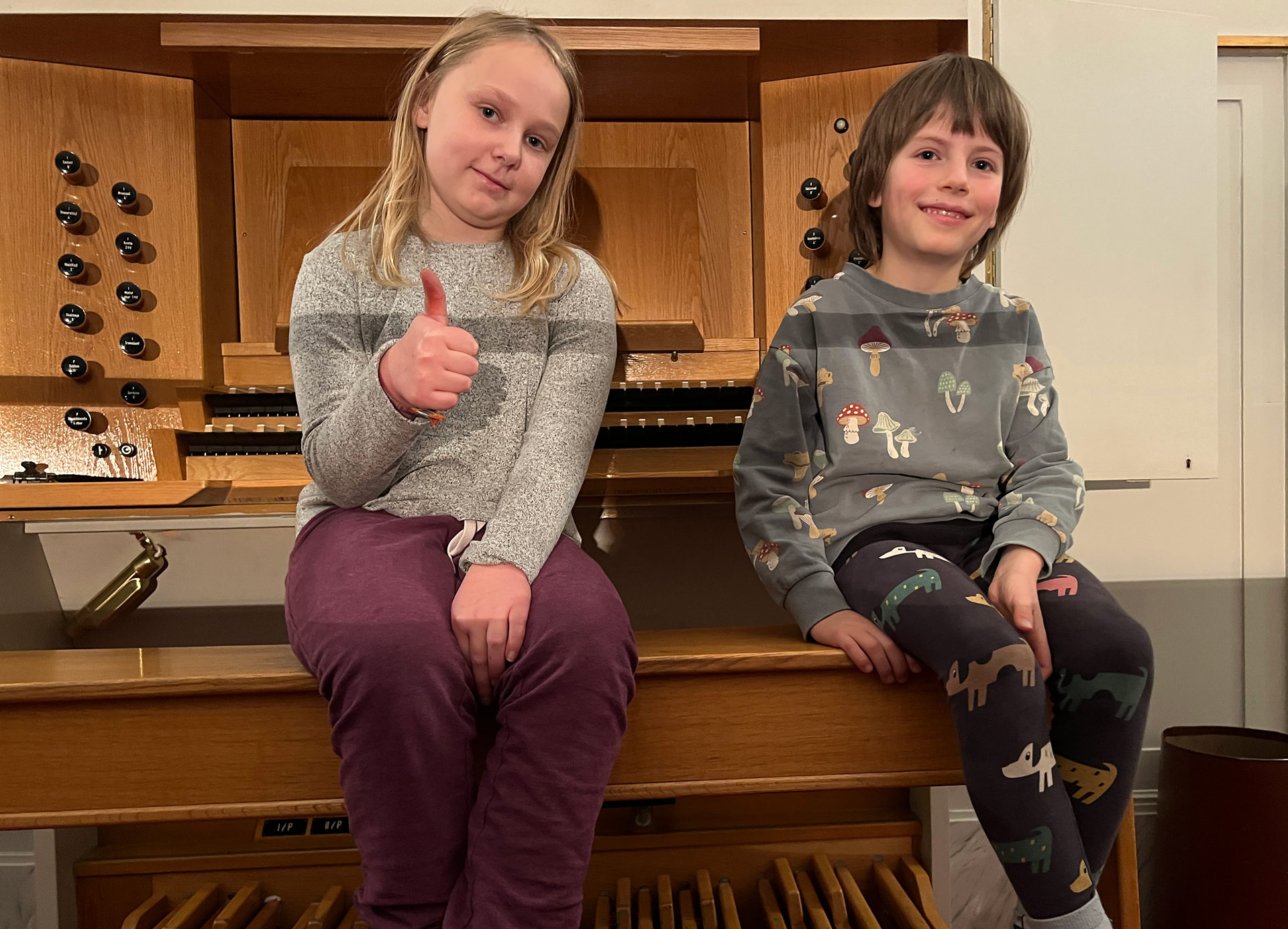 Två lågstadieelever sitter vid en kyrkorgel. Båda ler, en gör tummen upp.
