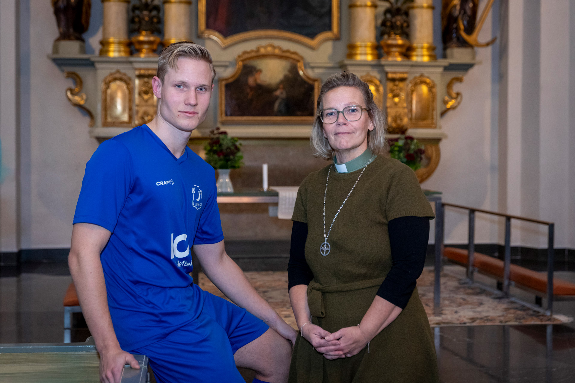En ung man i blåvita fotbollskläder samt en kvinnlig diakon  står framför altartavla.