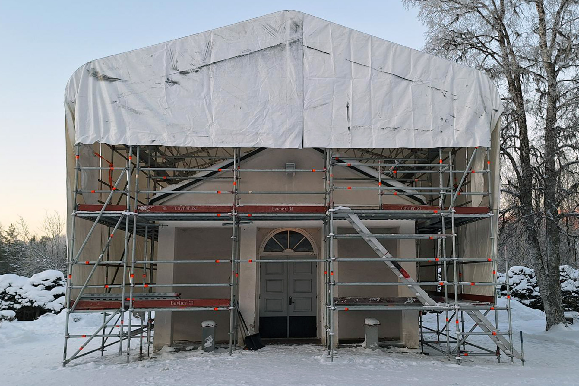 Ett litet vitputsat kapelll intäkt i byggnadsställning och vit presenning.