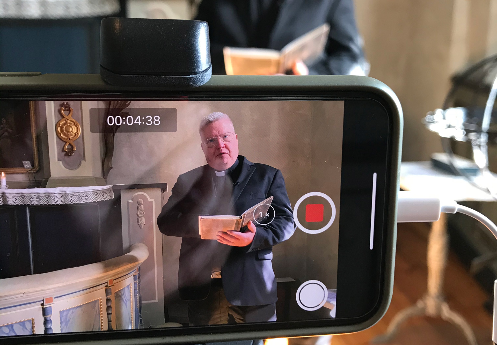 Genom skärmen på en mobiltelefon ser vi en präst som håller en andakt. 