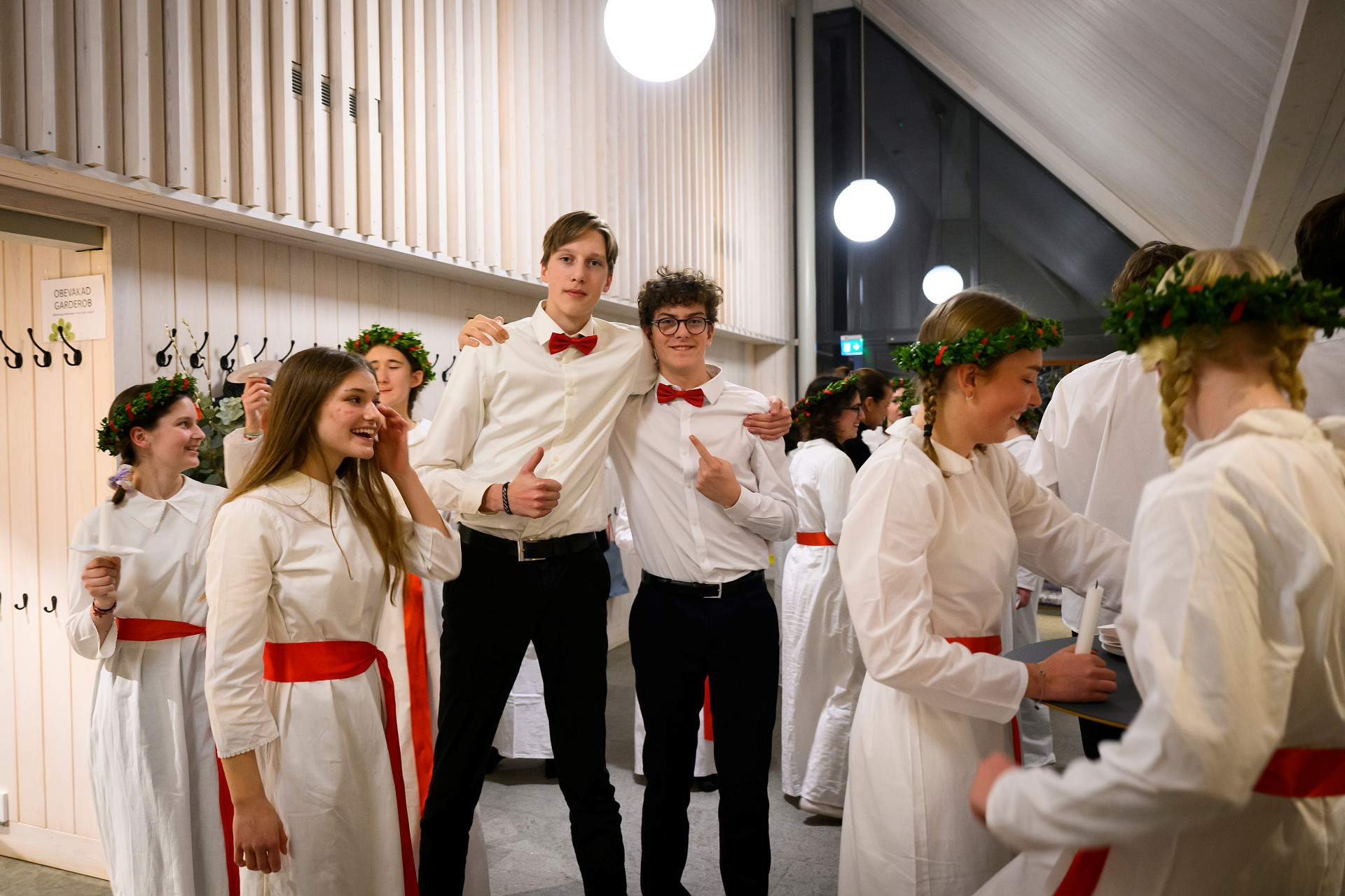 Flera ungdomar står luciaklädda i kyrkan. Två finklädda unga män håller om varandra och gör tummen upp mot kameran.