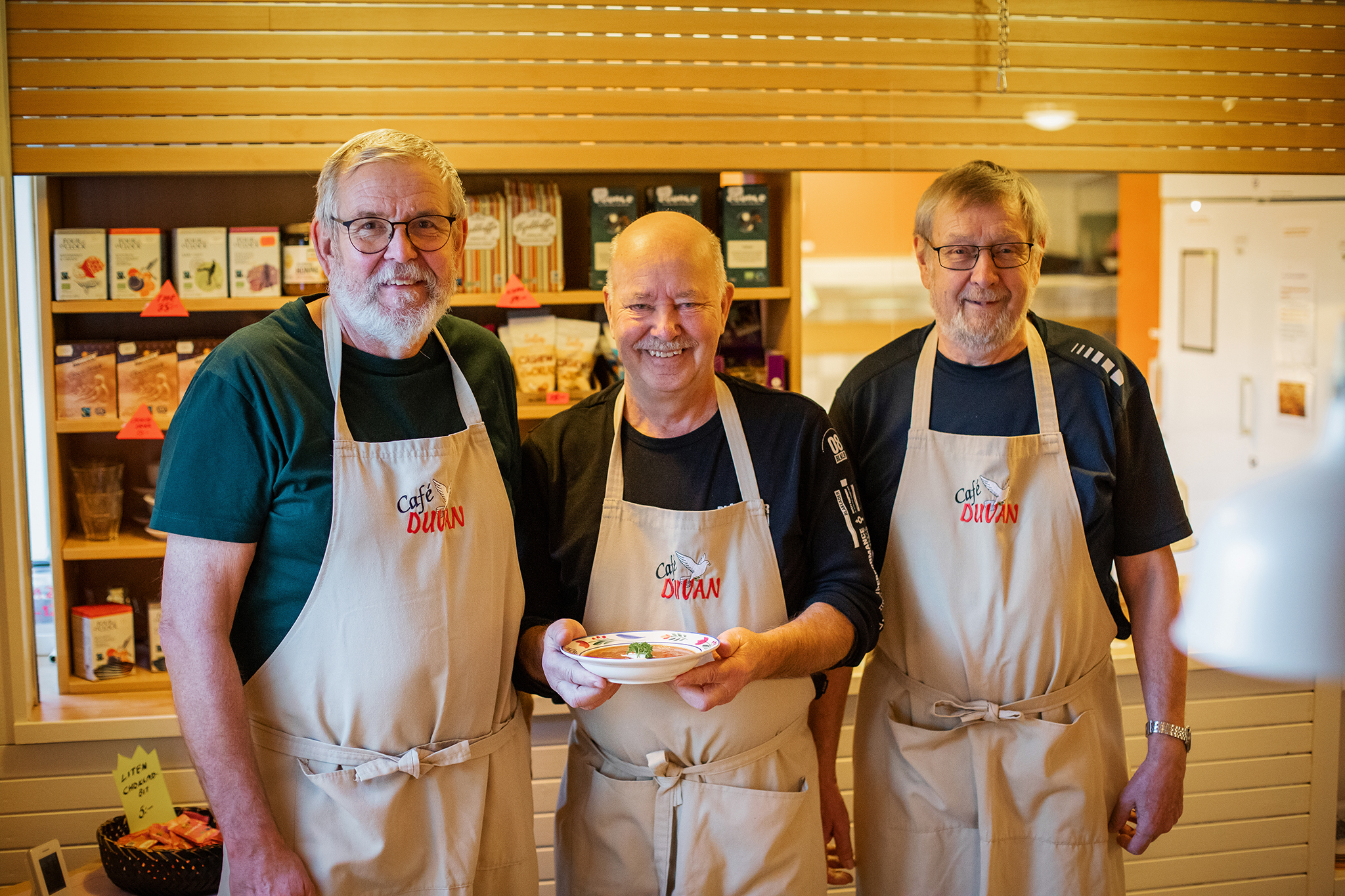 Tre män med förkläden i cafémiljö. Mannen i mitten håller i en tallrik med soppa. 