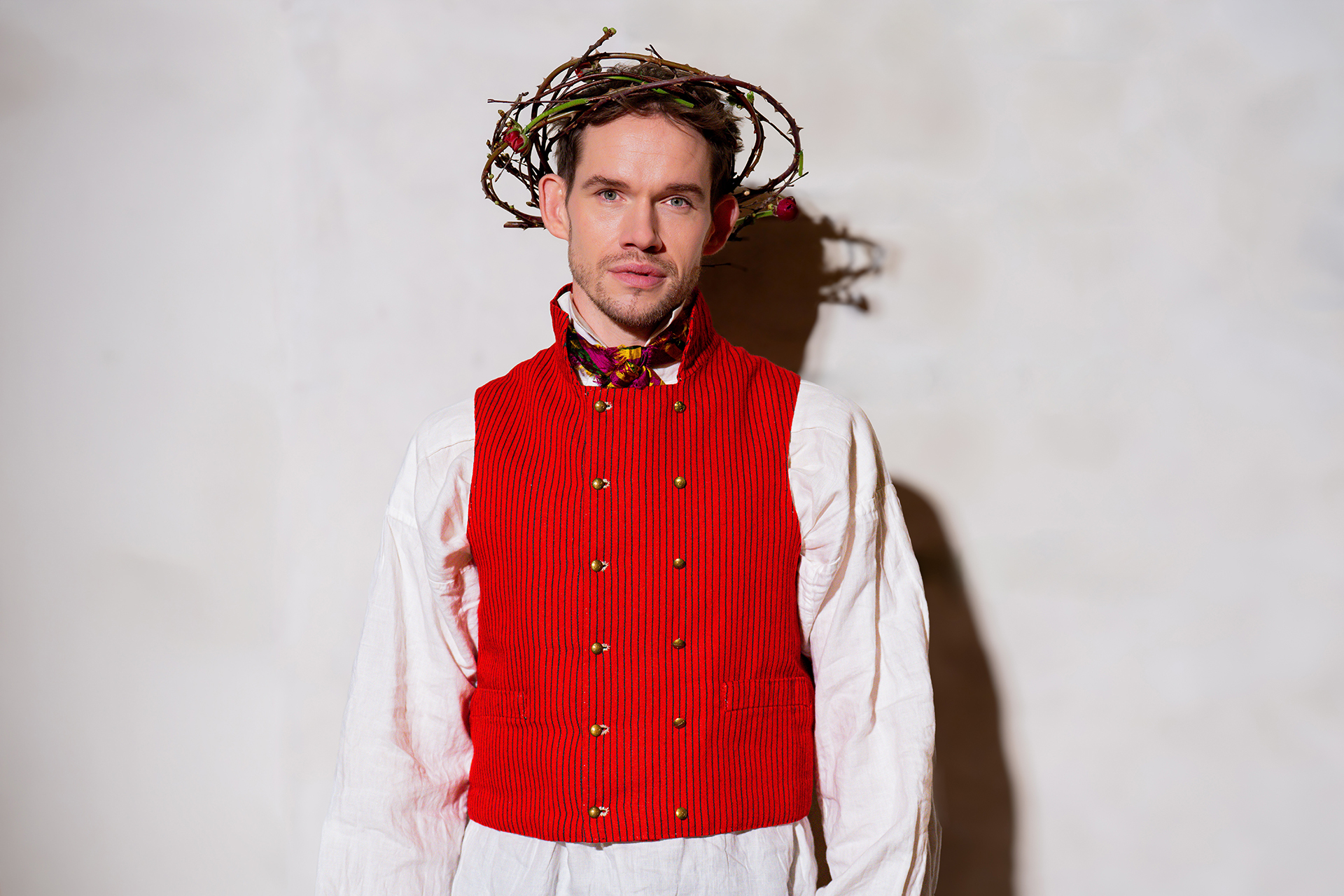 Sångaren David Alvefors mot vit bakgrund med folkdräkt i vitt och rött samt en törnekrona istället för blomsterkrans på huvudet.