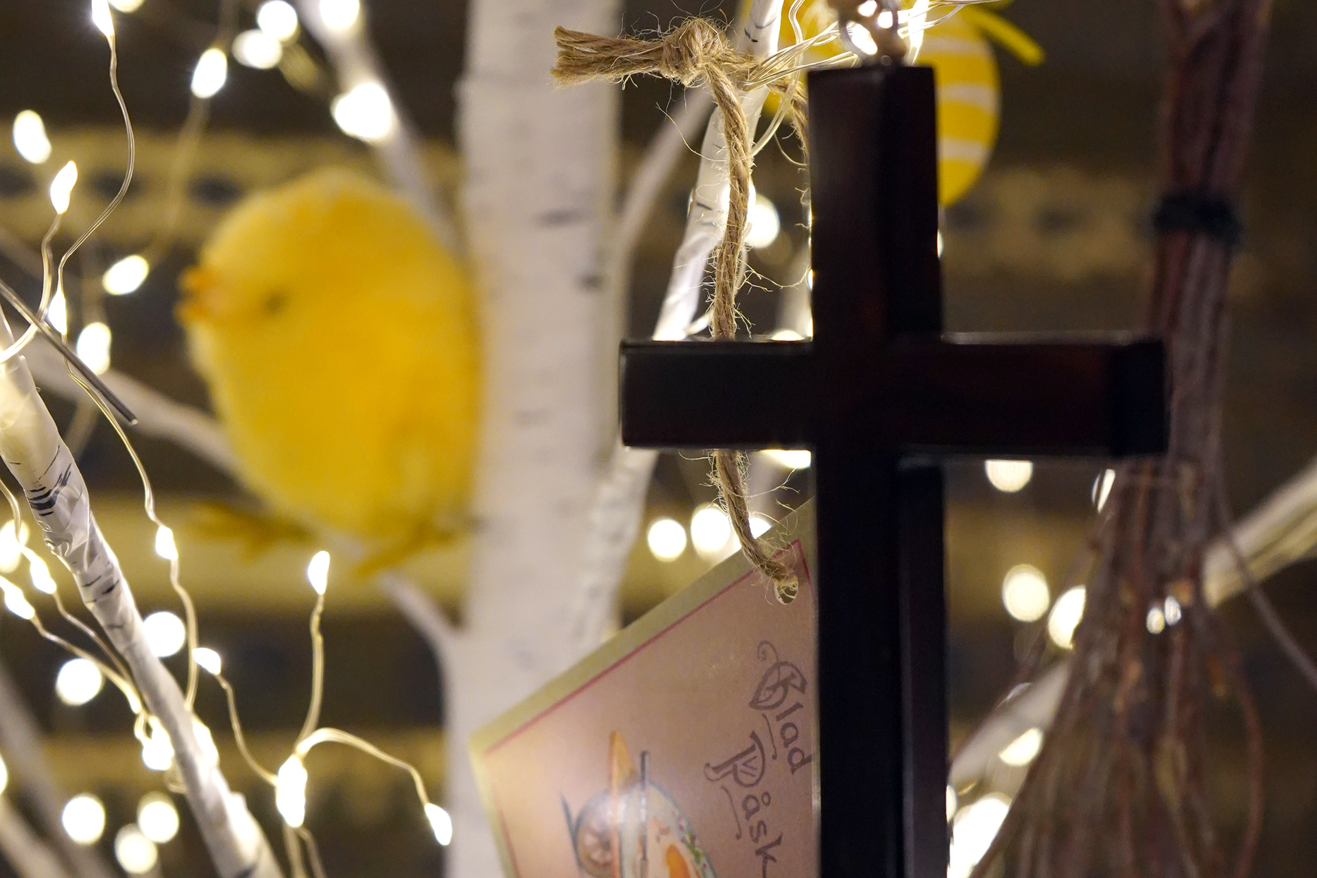 Ett kors, ett påskkort och en gul kyckling syns mot bakgrund av glimmande ljusslingor och en björkstam i ett påskbetonat arrangemang.