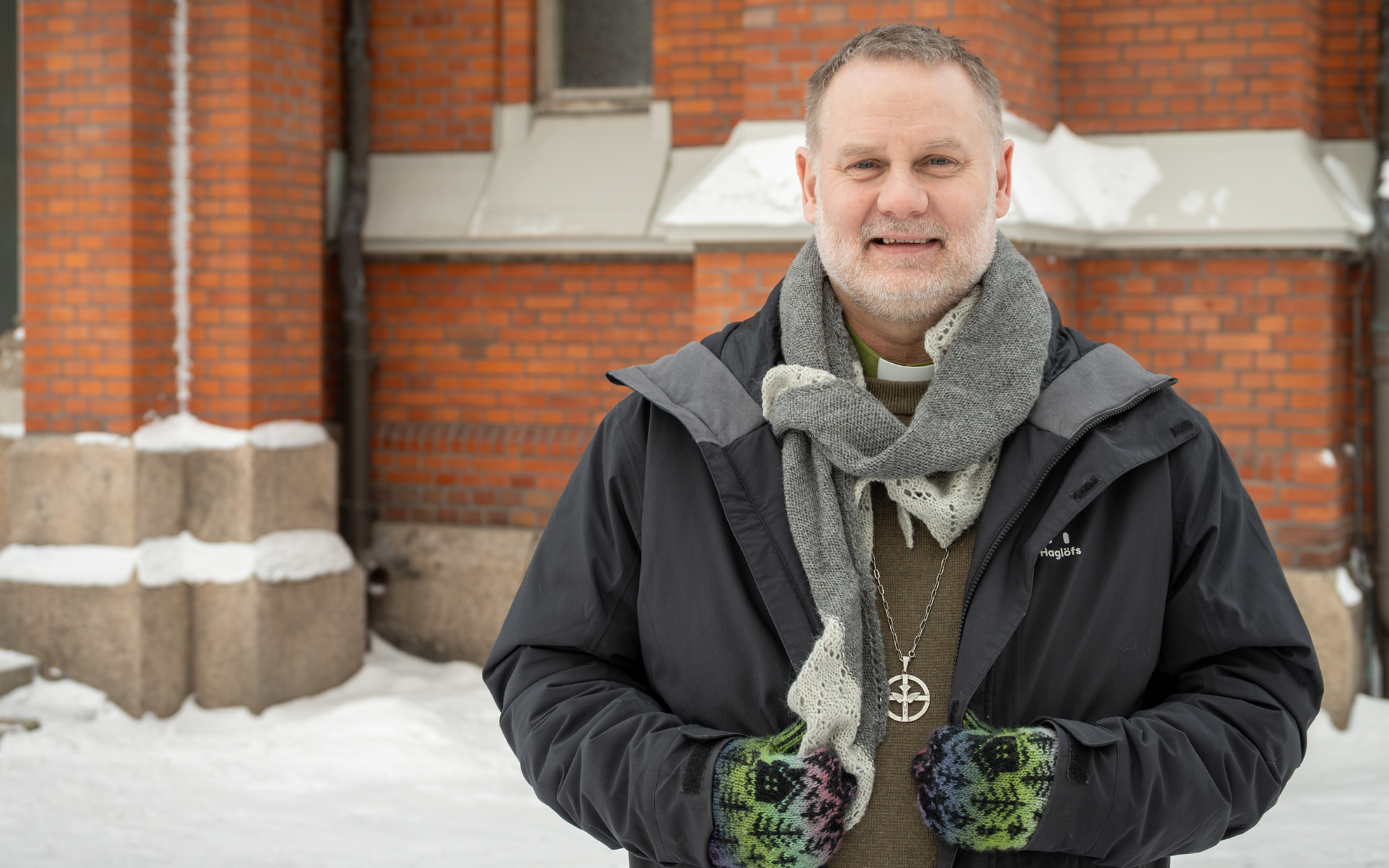 Man iklädd vinterjacka, stickad halsduk, stickade vantar framför röd tegelvägg