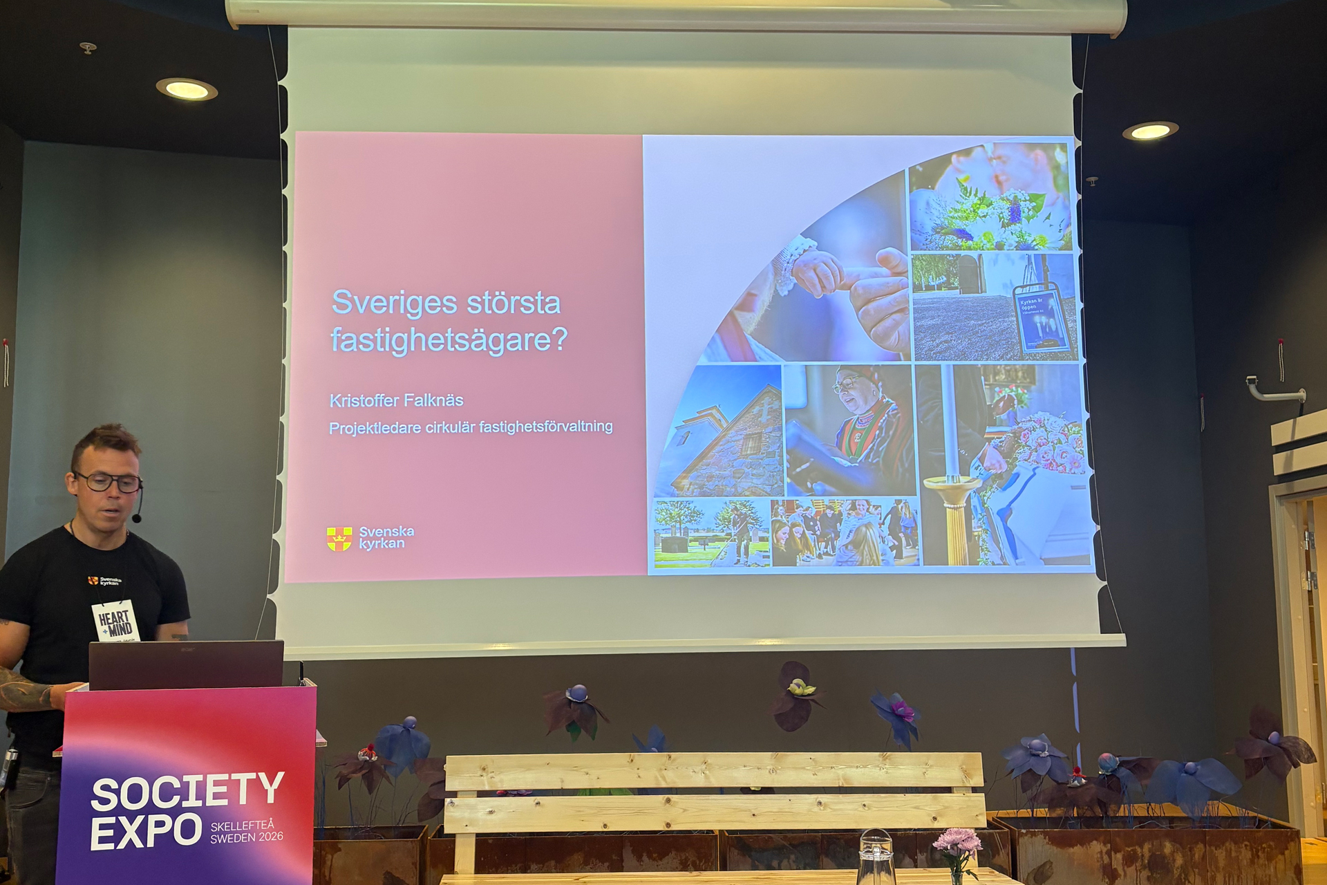 En person står vid en talarstol bredvid en projektorduk och visar en presentation.