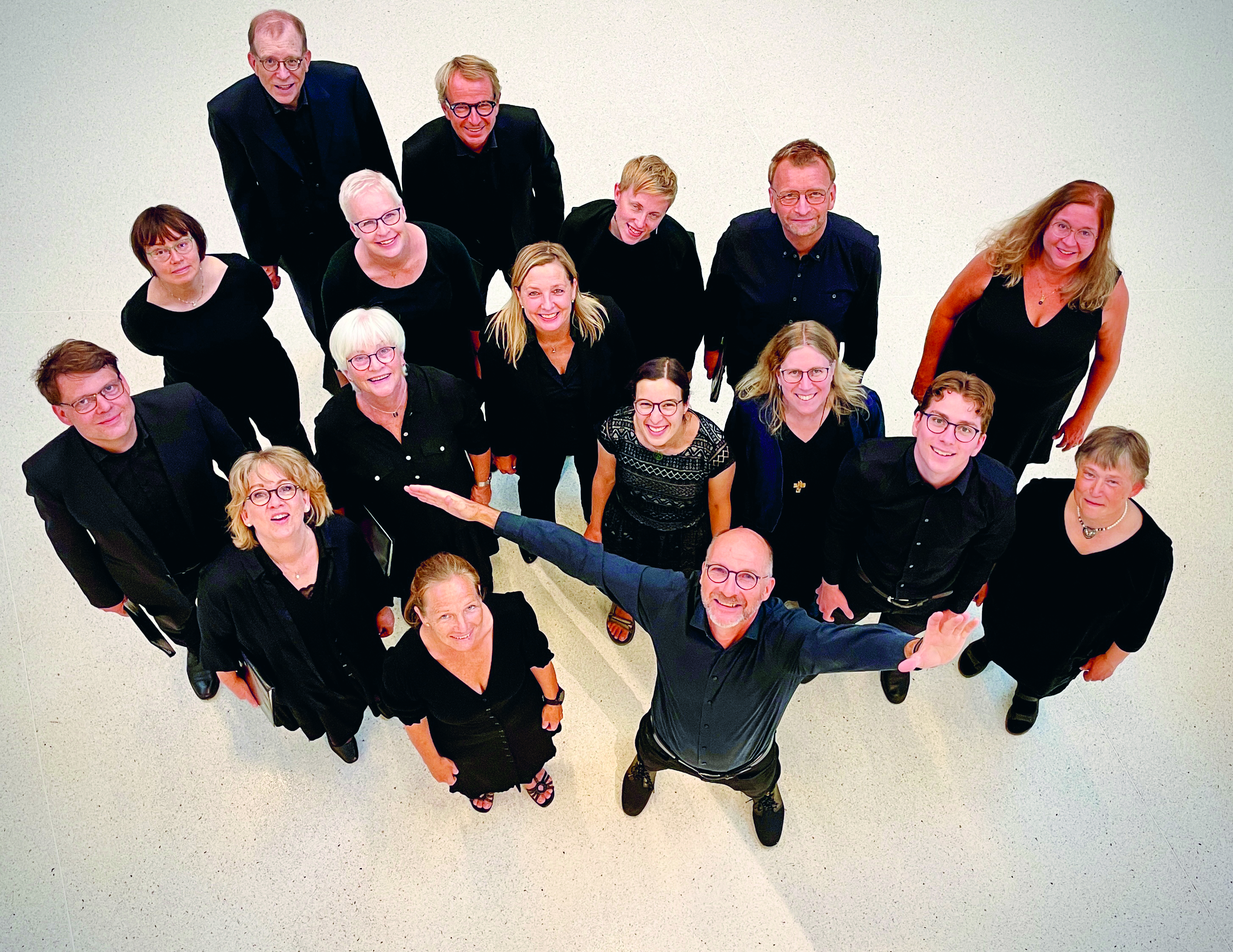 17 kvinnor och män klädda i svart står på ett vitt golv och tittar upp mot fotografen.