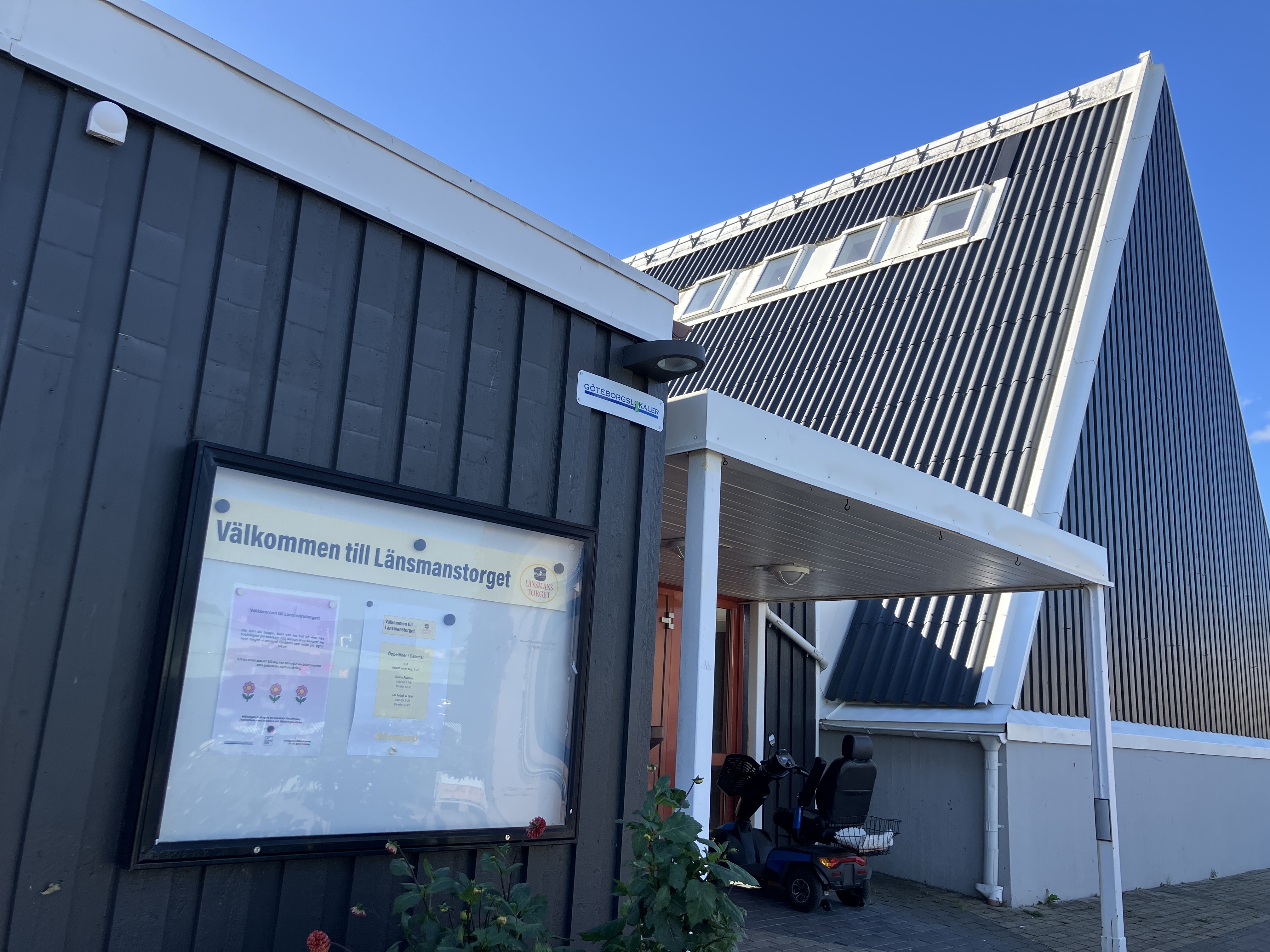 Närbild på en grå träbyggnad, som tidigare var en kyrka. På väggen sitter en anslagstavla med rubriken "Välkomment till  Länsmanstorget" och en skylt med GöteborgsLokalers logga. Vid ingången till byggnaden står en promenadscooter.