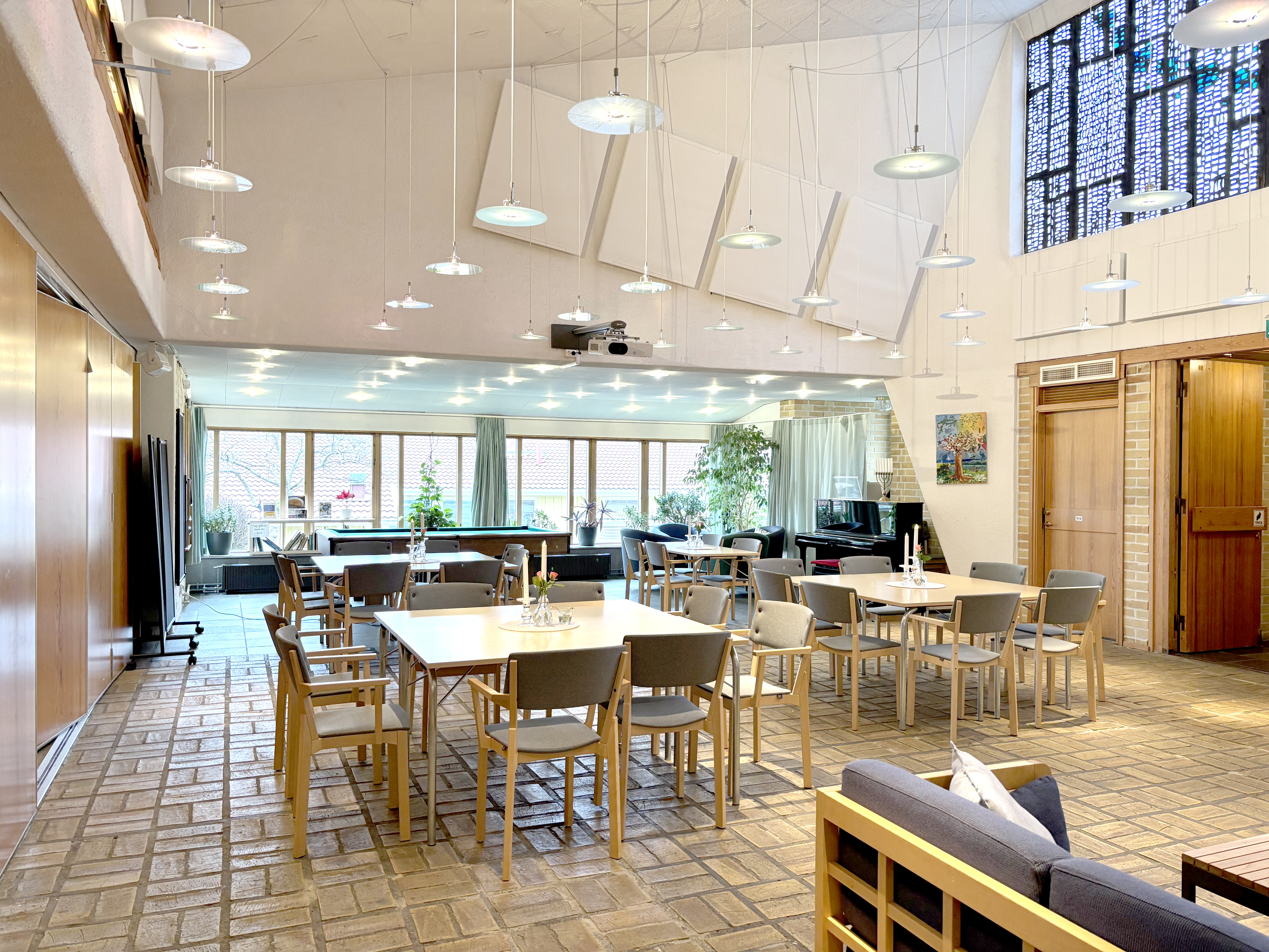 En lokal möblerad med i huvudsak stolar och bord. Högt i tak, med nedpendlad belysning. Golv i gult tegel, till vänster vikvägg, i fonden stora fönsterpartier, till höger entrédörrar.