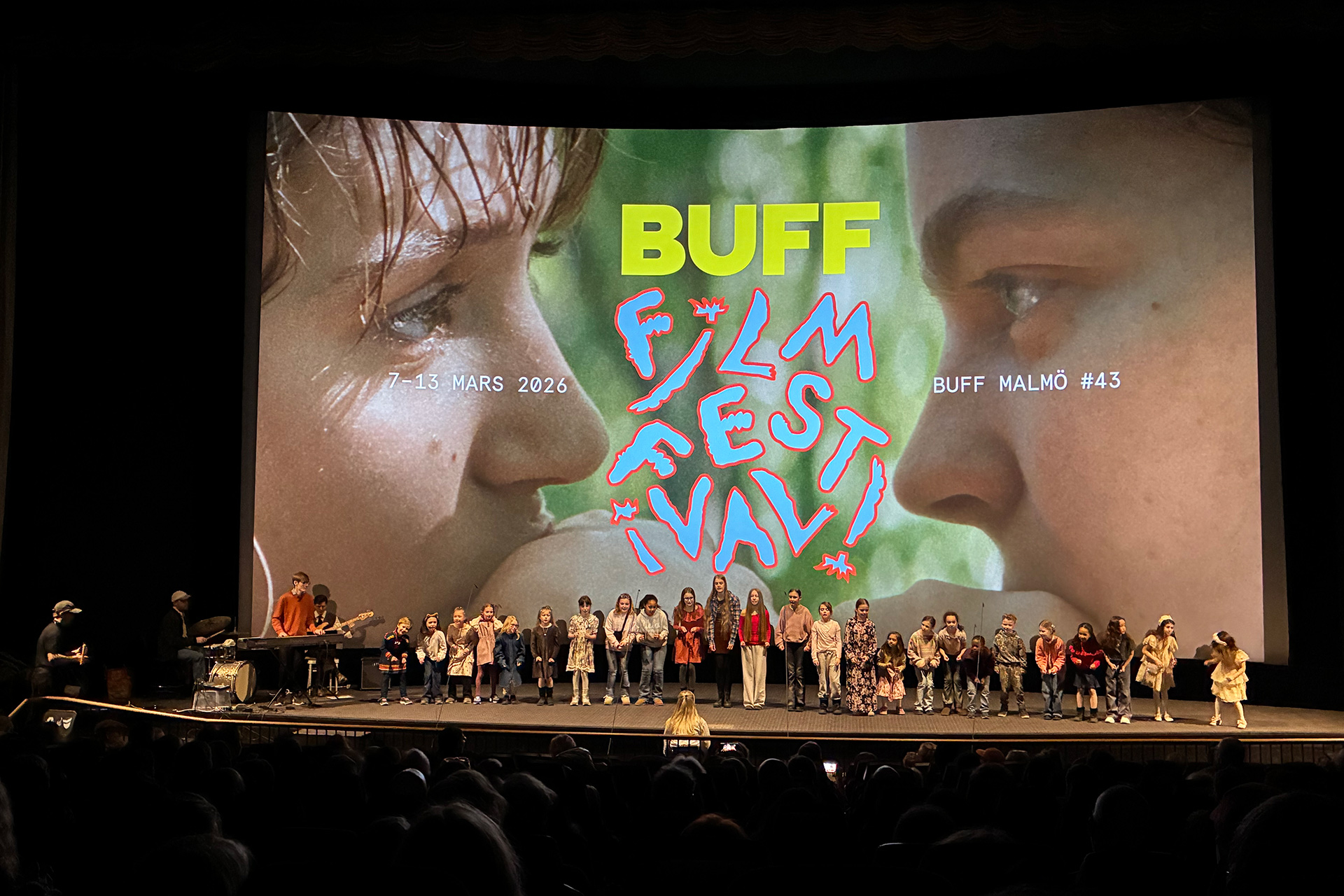 En barnkör står på en scen i en biosalong. På scenen finns också en trummis, en som spelar keyboard och en basist. Bakom kören, på filmduken, syns en närbild av två tonåringars ansikten i profil. De tittar på varandra. Mellan ansikten står texten BUFF filmfestival 7–13 mars 2026, BUFF Malmö #43.