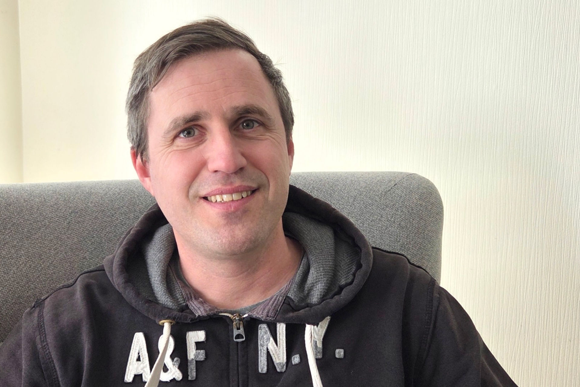 En man med grått kort hår är klädd i en mörk luvtröja med vit text framtill. Han sitter i en fåtölj. 