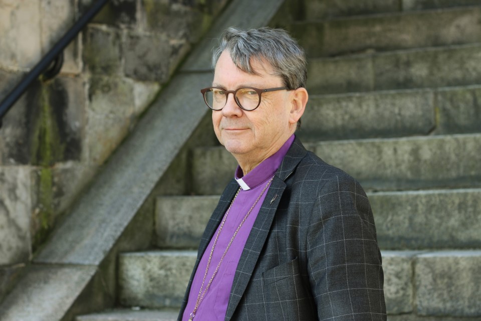 En man med glasögon och grånat, klädd i lila prästskjorta och mörk kavaj. Runt halsen hänger en guldkedja. Han står i profil, intill en stentrappa utomhus.