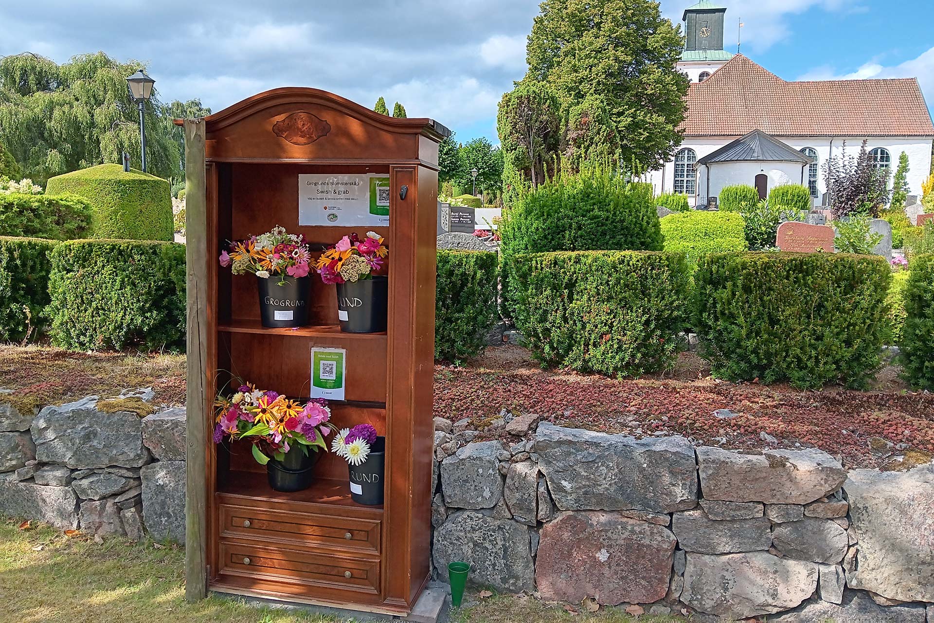 Invid kyrkomuren står ett skåp i trä. I skåpet står bundna buketter i spannar.