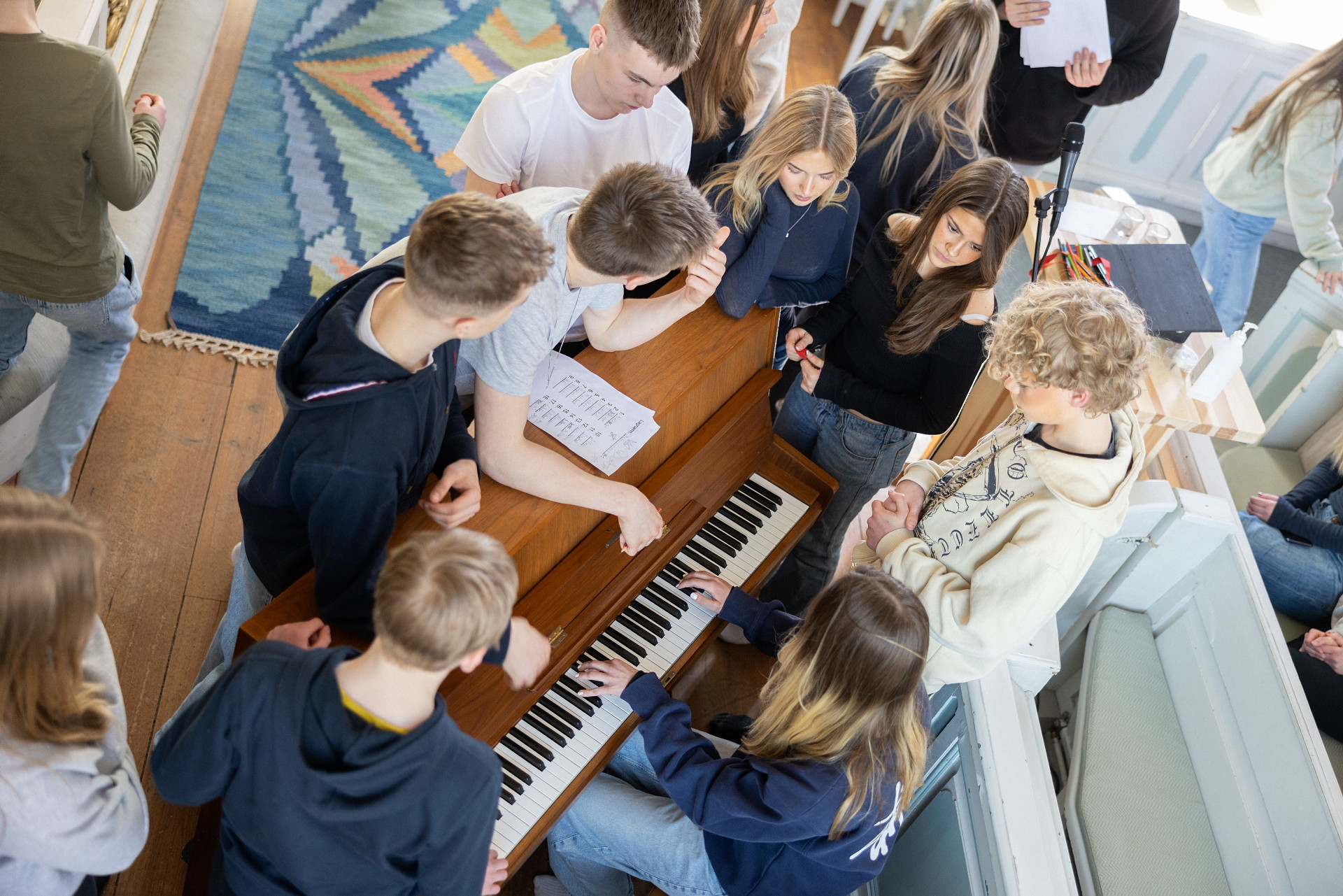 En grupp unga människor står runt ett piano och snackar.
