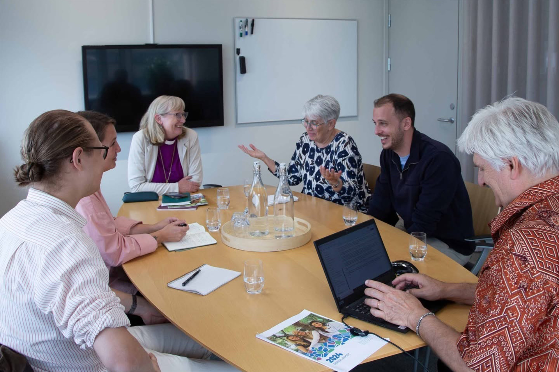 En grupp människor pratar, antecknar och skrattar runt ett bord i ett konferensrum. 