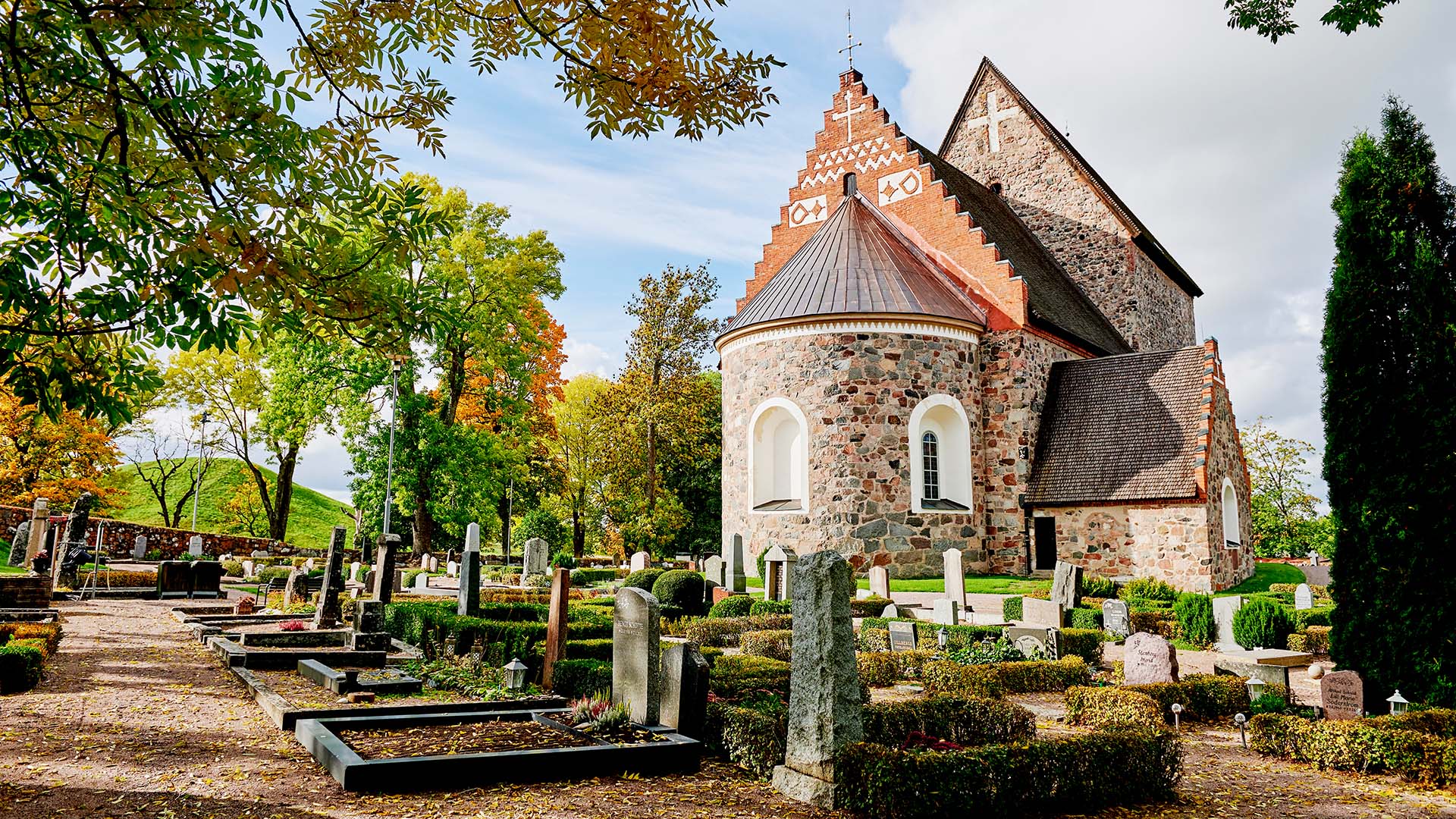 En kyrka i solsken. Framför kyrkobyggnaden syns en kyrkogård.