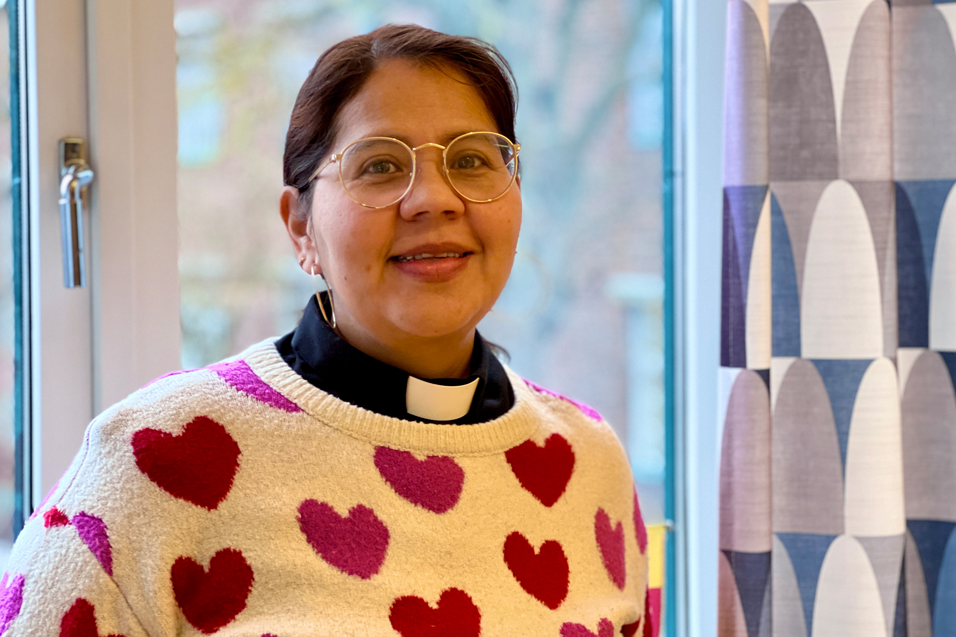 En leende kvinna med prästkrage och tröja med hjärtan på står framför ett fönster. 
