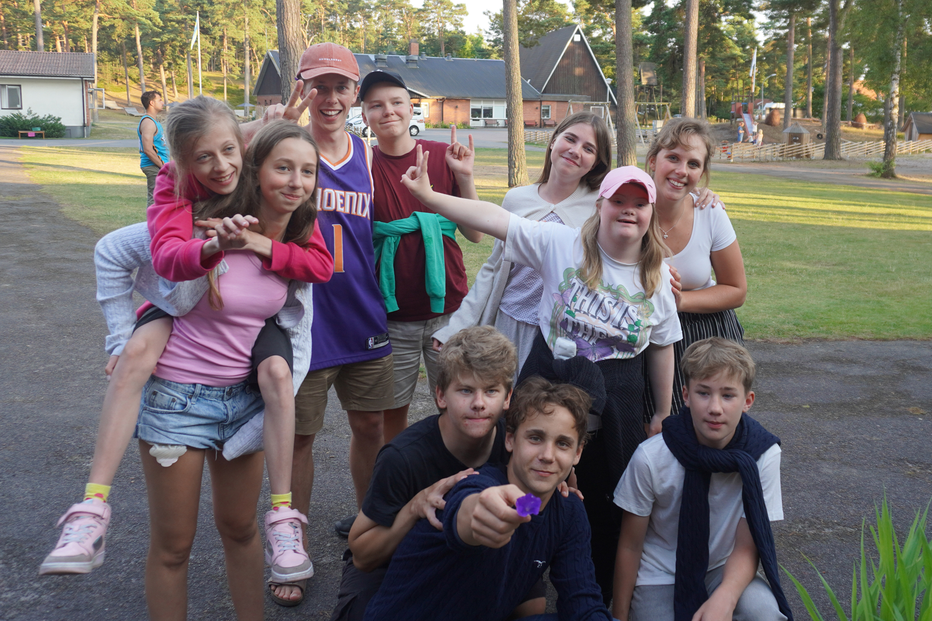 Tio glada ungdomar står tillsammans utomhus. En person rider på en annan rygg. Det är sommar 