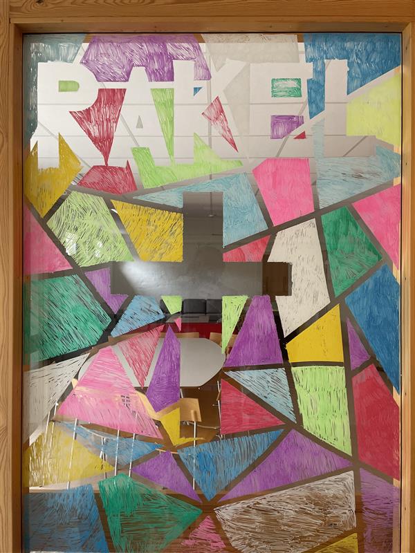 Ett fönster som är fyllt med färgat silkespapper i olika färger, där glaset är genomskinligt bildar tomrummet ordet Rakel, samt ett kors.