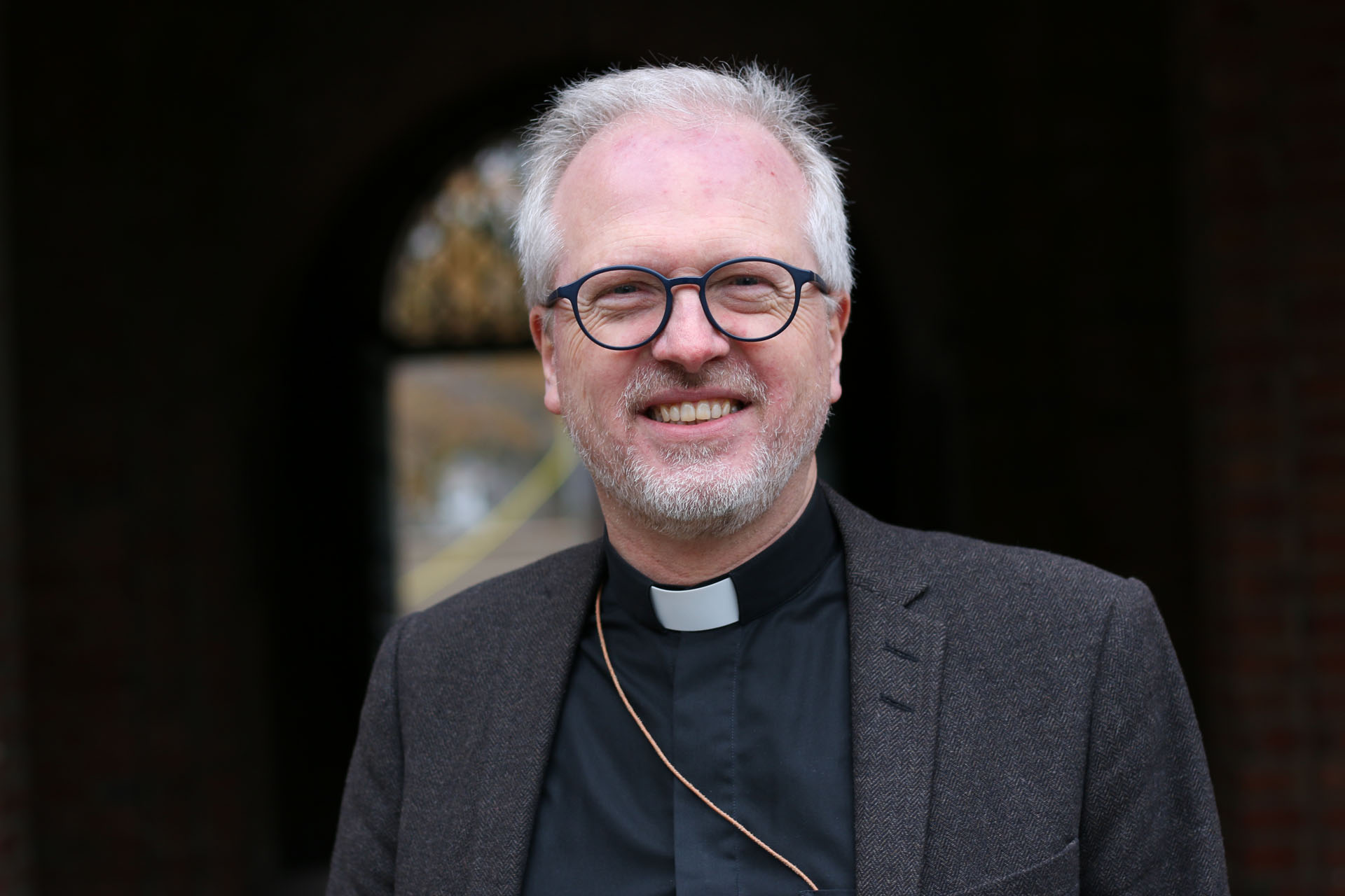 En leende man med prästkrage och glasögon tittar in i kameran.