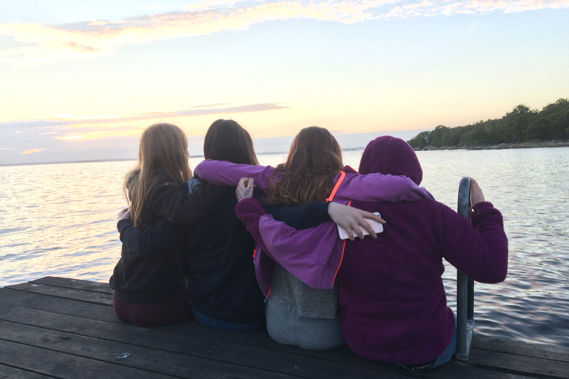 Fyra ungdomar sitter på en brygga och håller varandra om ryggarna. De tittar ut över en sjö i skymningsljus.