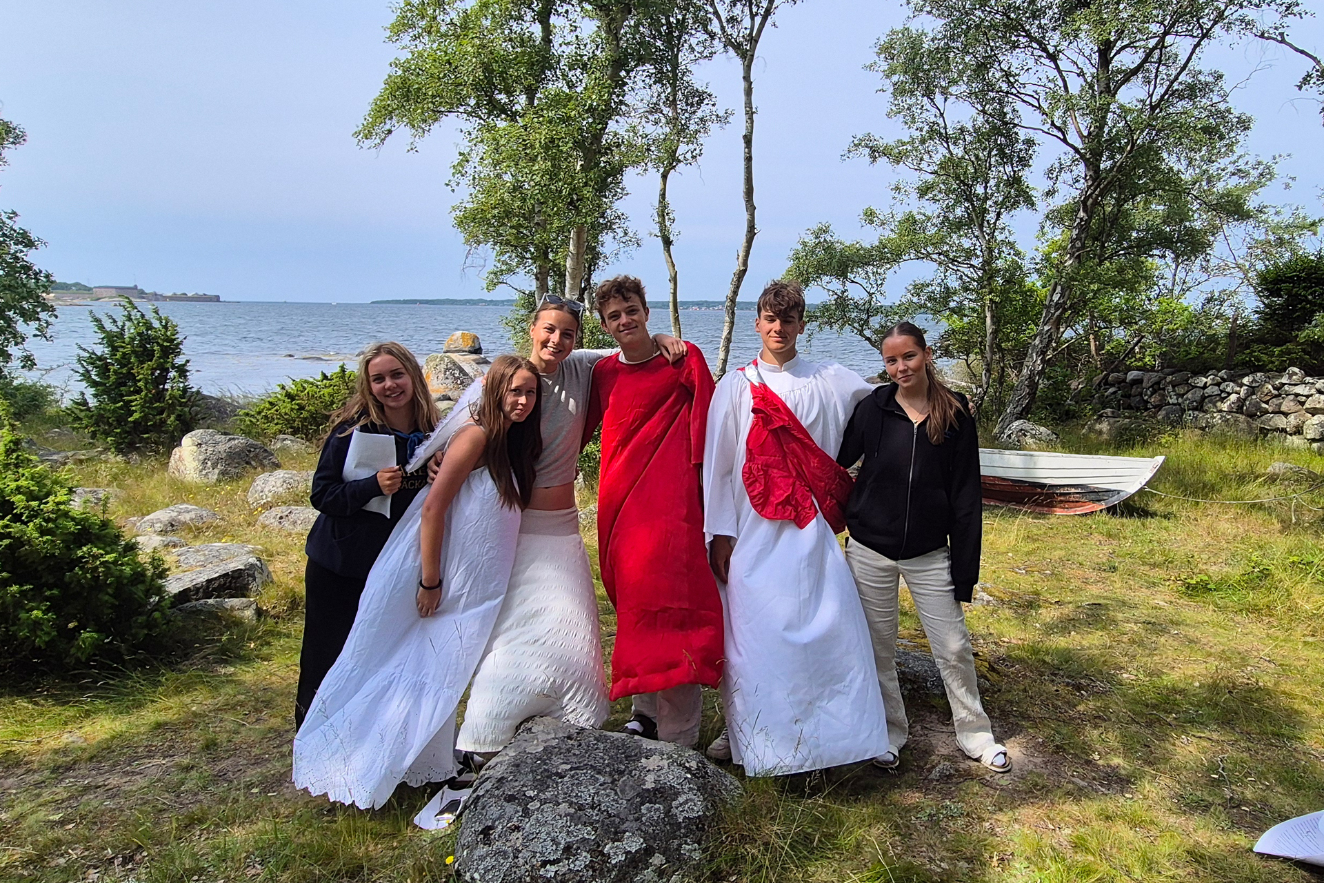 En grupp ungdomar står med armarna om varandra och tittar in i kameran. I bakgrunden syns en hav och en eka liggandes på land.