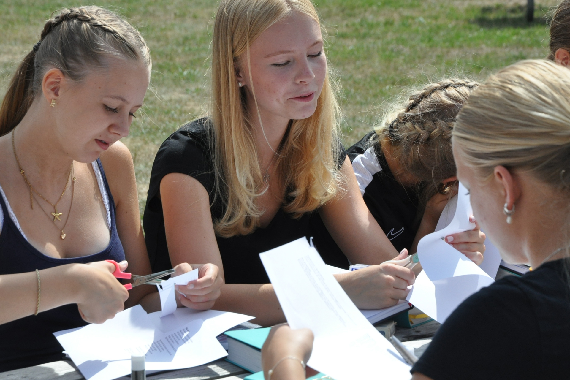 En grupp ungdomar klipper i papper vid ett bord utomhus. 
