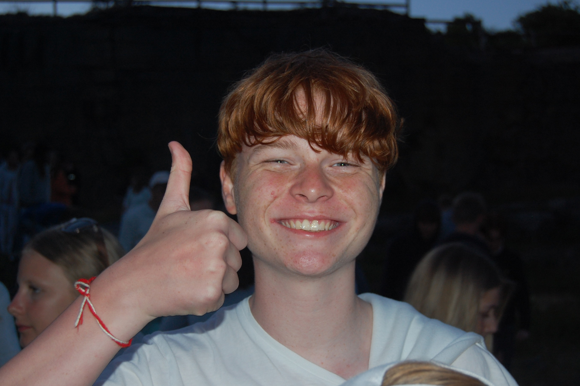 En glad kille gör tummen upp. I bakgrunden syns en grupp ungdomar. Det är kväll.