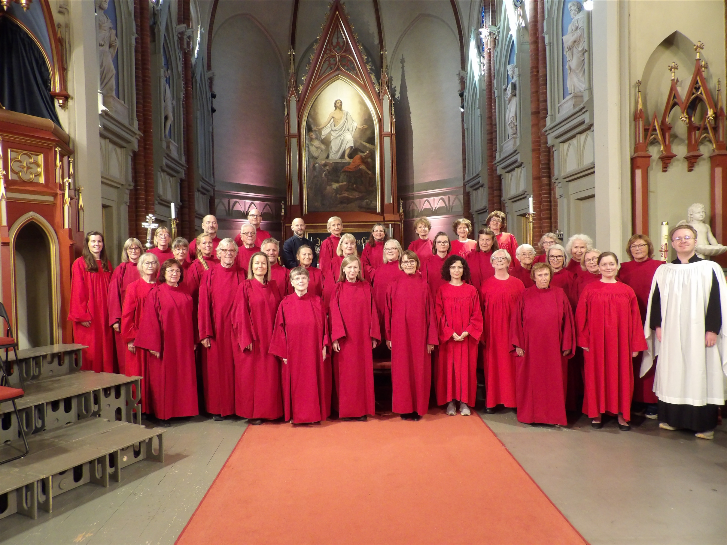 Stor kör klädd i röda kåpor står uppställd i mittgången i en kyrka, med höga valv och en altartavla föreställande en religiös scen bakom.