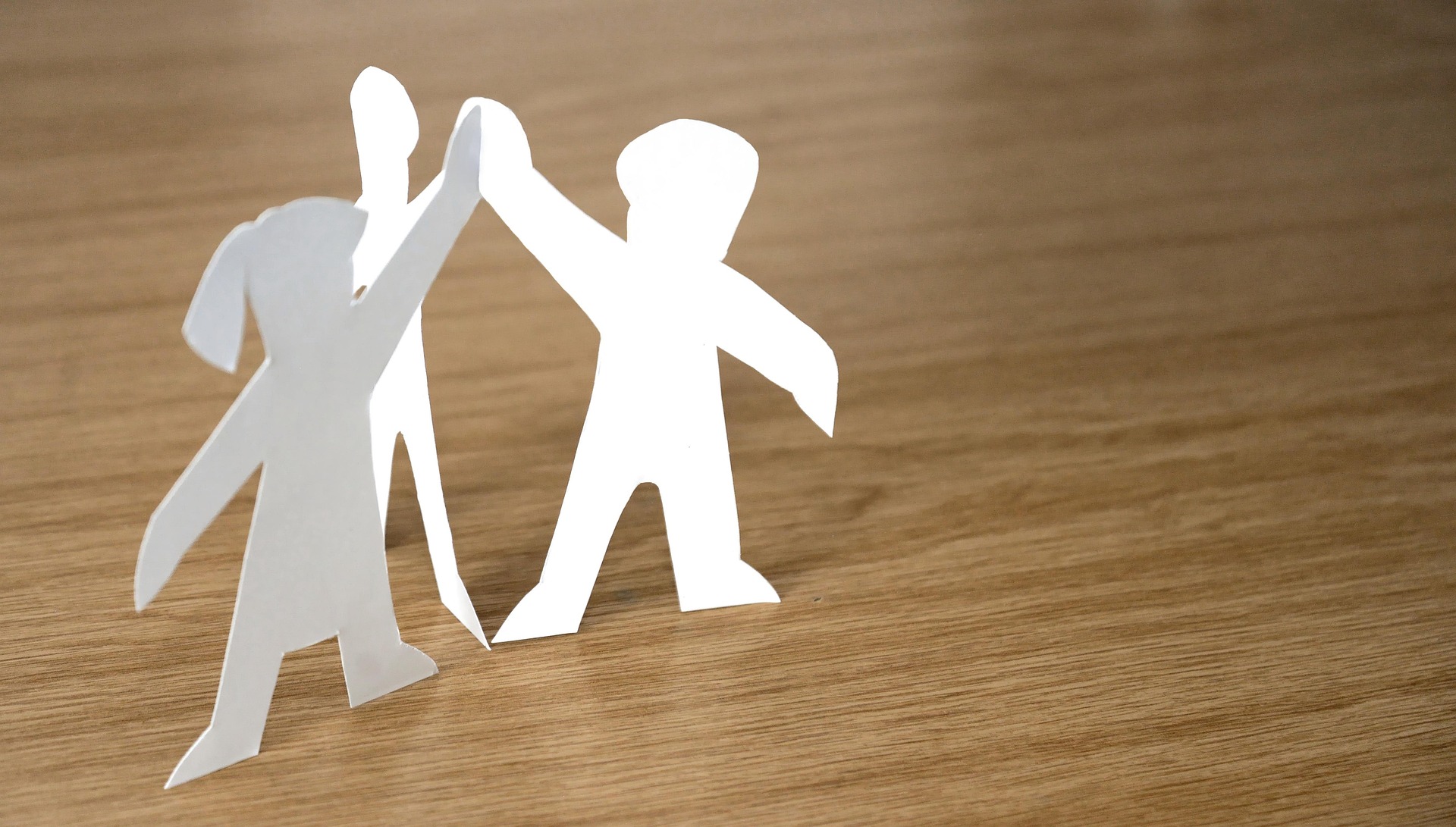 Tre urklippta pappersfigurer på en träskiva höjer händerna tillsammans. 