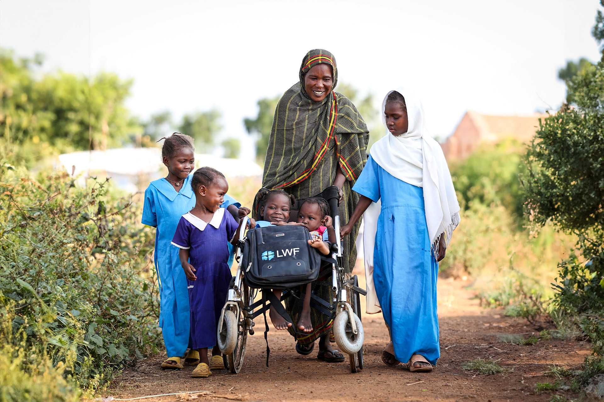 Två flickor sitter i en rullstol som skjutas av en kvinna, kring rullstolen går några barn. 