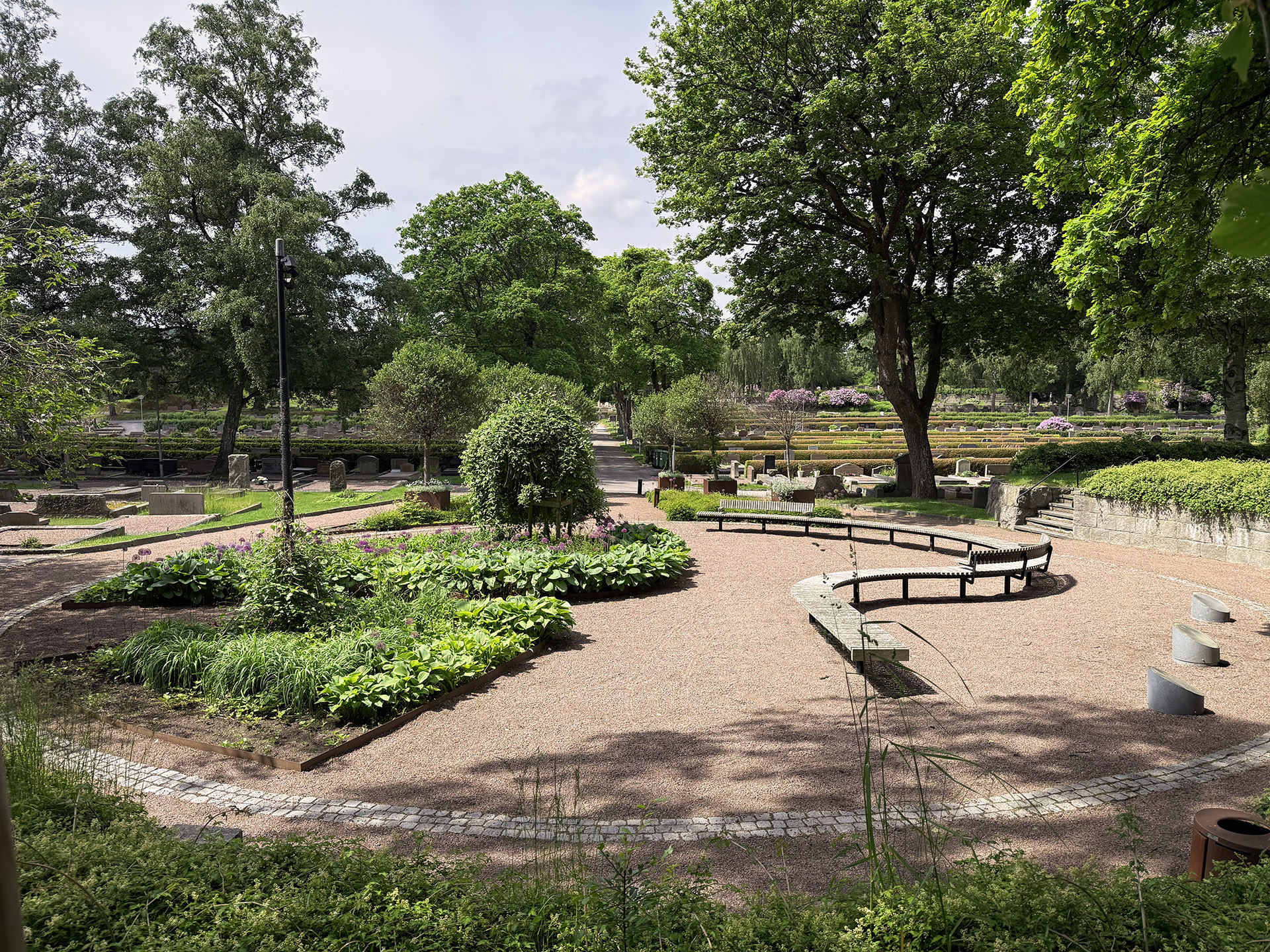 Grusad cirkelformad plats inramad av gatsten på en kyrkogård. Lummig grönska i form av träd och buskar samt perenner i planteringar. En sittbänk som slingrar sig utmed högerkanten.