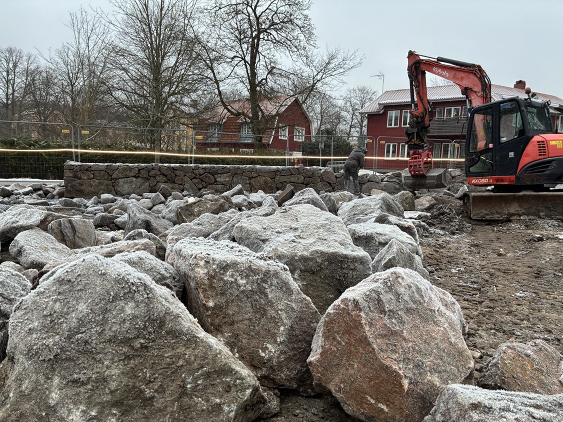 Grävmaskin står bland stora stenar, träd i bakgrunden.