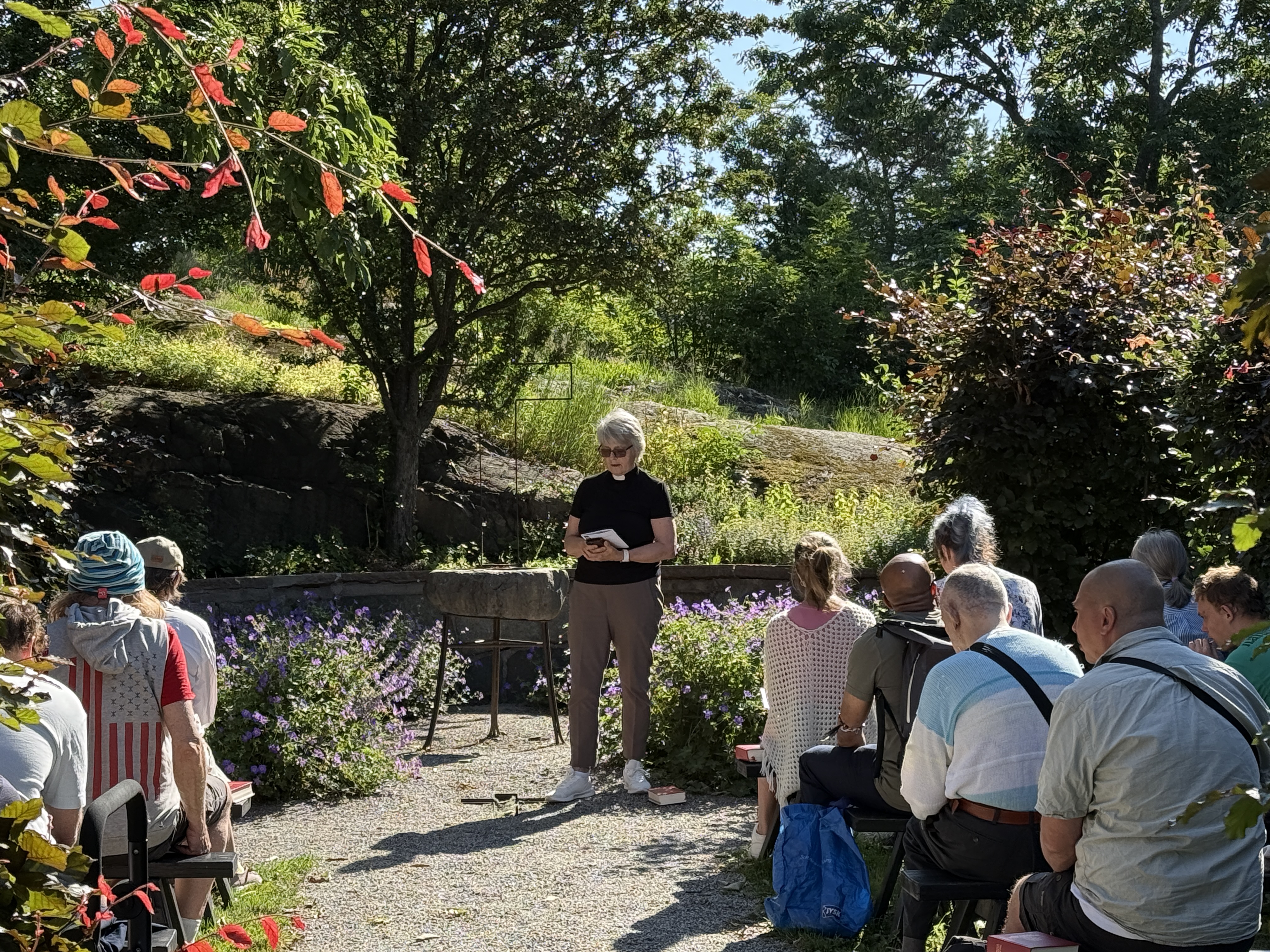 En grupp människor sitter på bänkar utomhus i en grönskande trädgård och lyssnar till en person som står på en grusgång och håller i en bok eller anteckningsblock. Lila blommor kantar gången och stora träd ger ett växelspel av sol och skugga. Röda och gröna blad ramar in bilden i förgrunden, medan klippor, buskar och frodig vegetation syns i bakgrunden under en klar och solig dag.