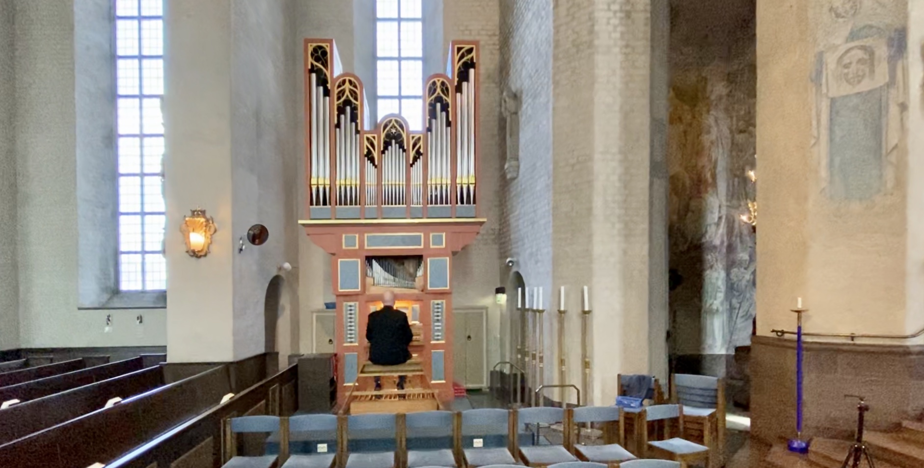 En orgel och en person som sitter vid den. kyrkbänkar, ljus, höga fönster. 